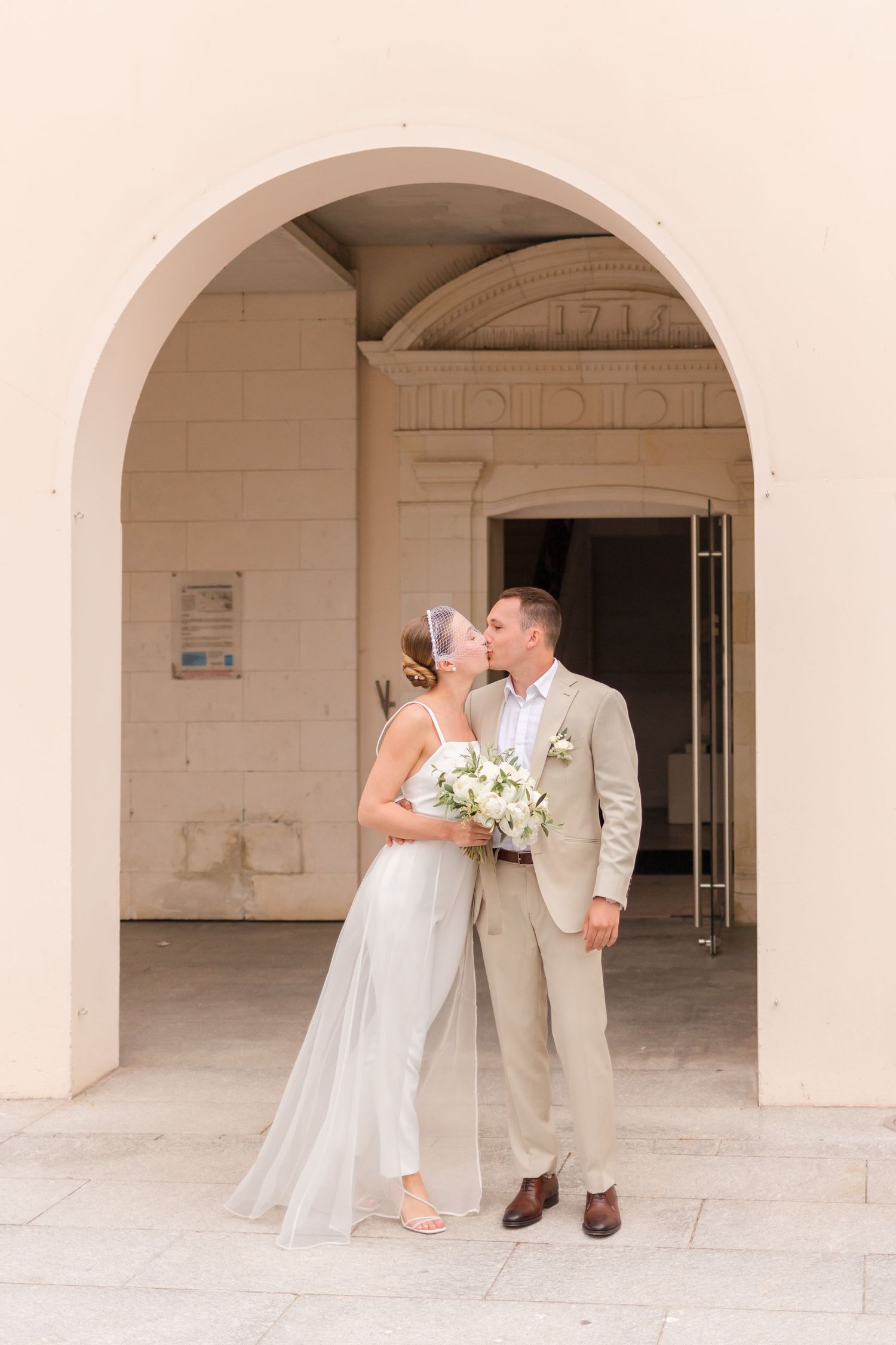 A romantic moment captured under an arched doorway with white flowers and elegant attire.