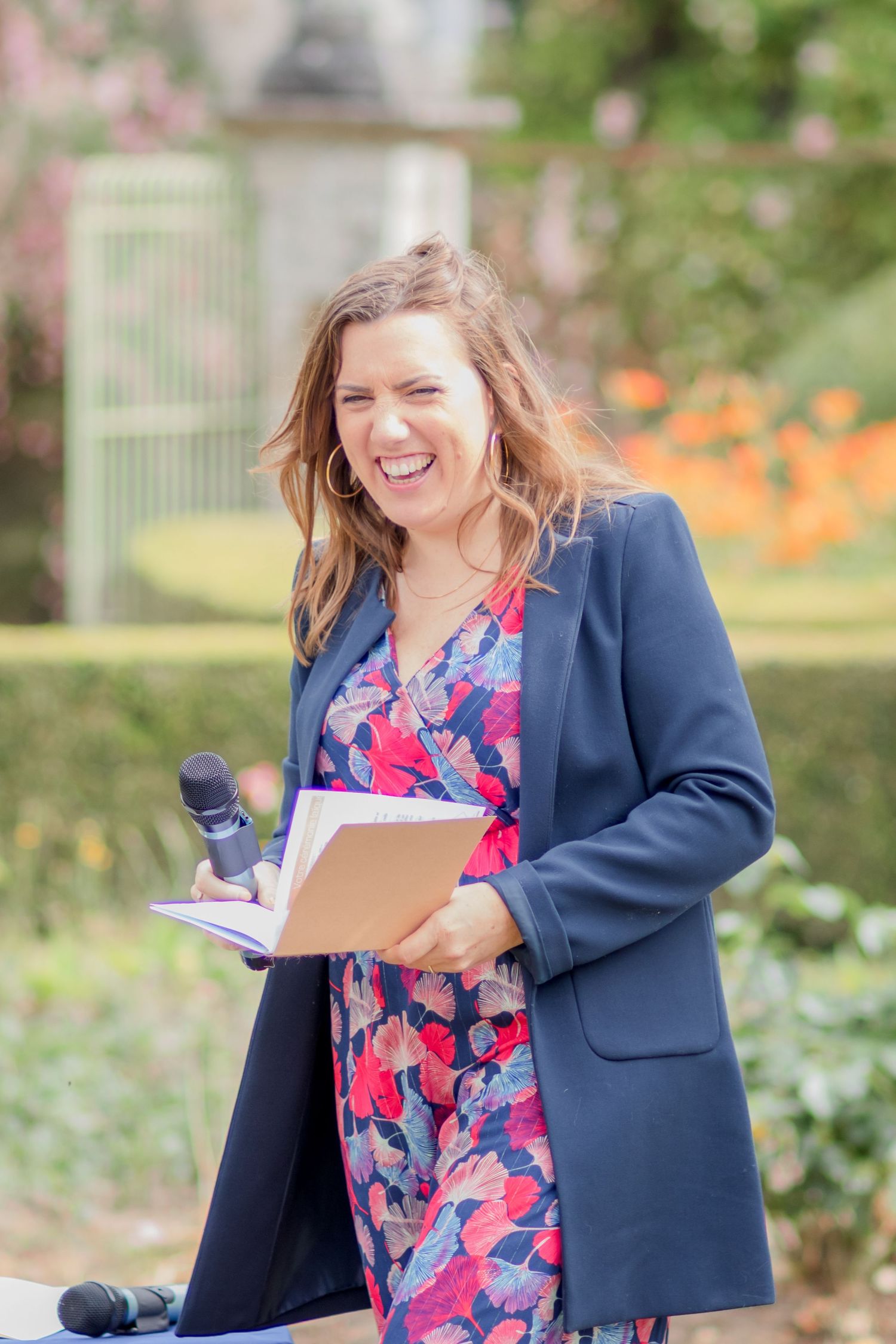 Woman in navy blazer and floral dress speaks at outdoor garden event.