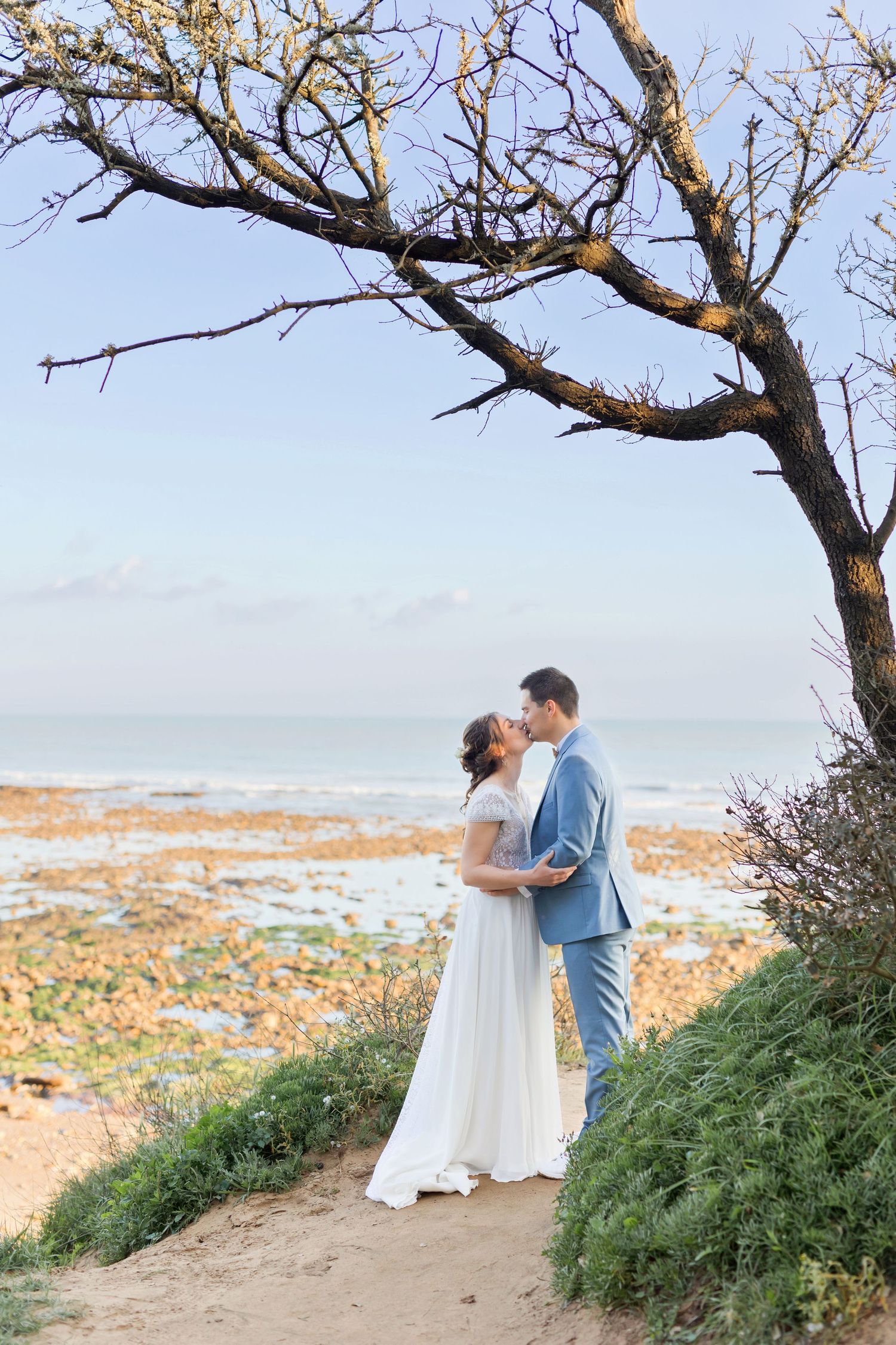 A coastal wedding photo with a couple embracing under a bare tree branch overlooking the beach.