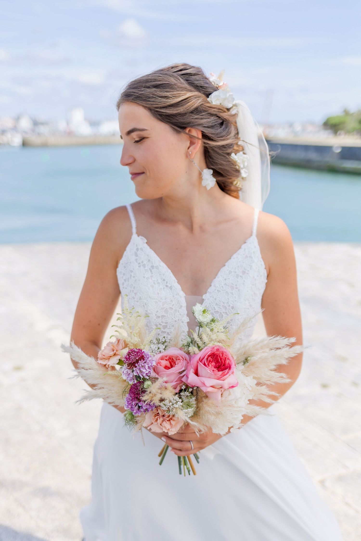 Elegant bride in a lace wedding dress holds a beautiful bouquet of pink roses and purple flowers by the water.