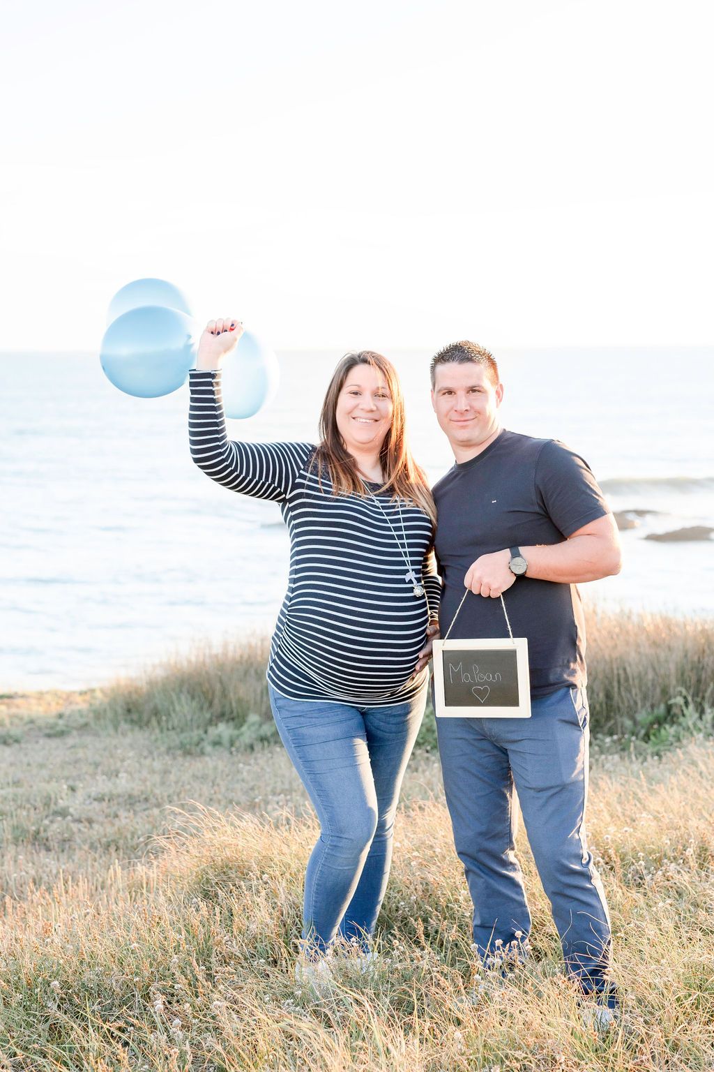 Expectant couple poses on coastal grassland with blue balloons and ultrasound photo during maternity shoot.