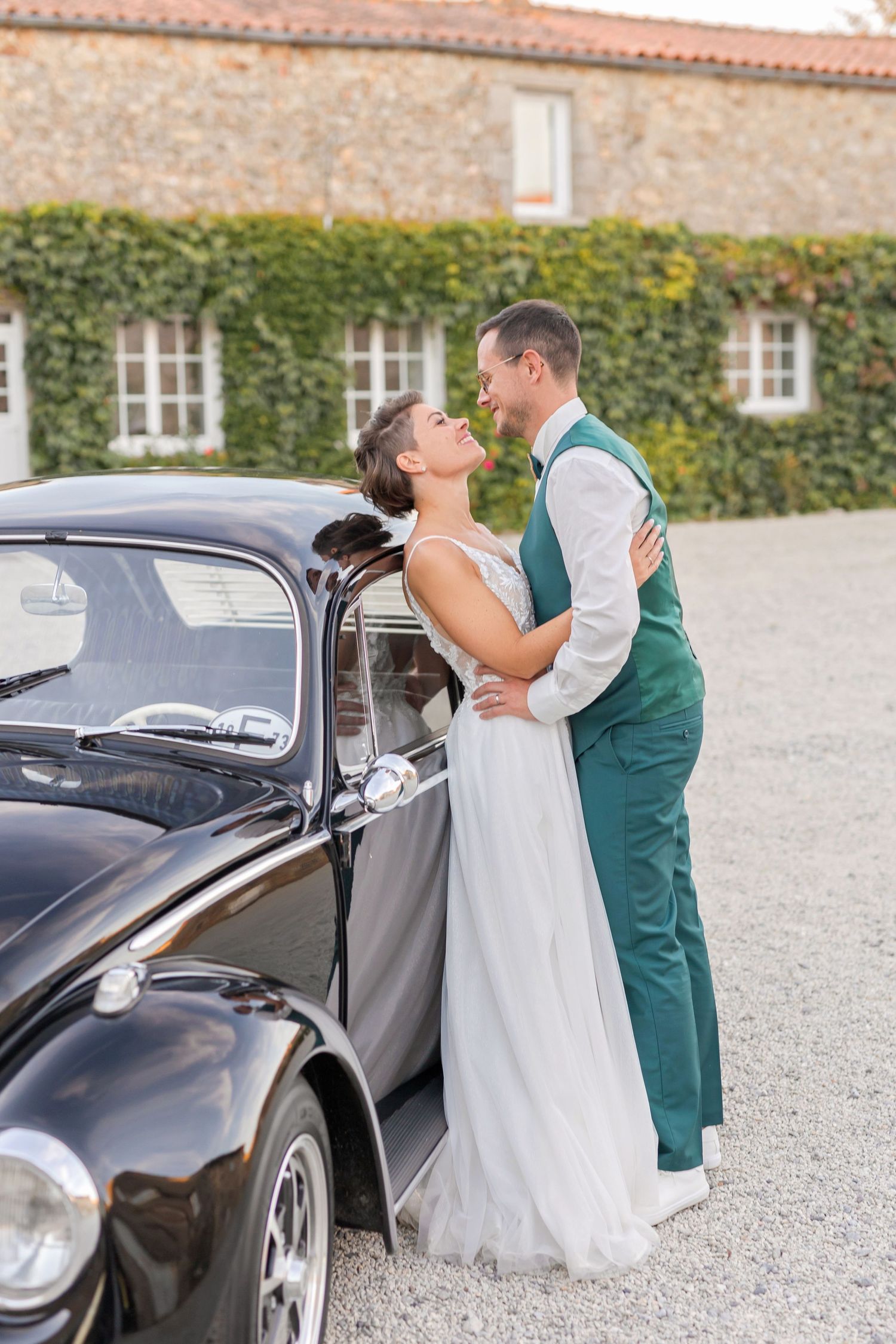 Wedding couple poses romantically against classic black Volkswagen Beetle near ivy-covered building.