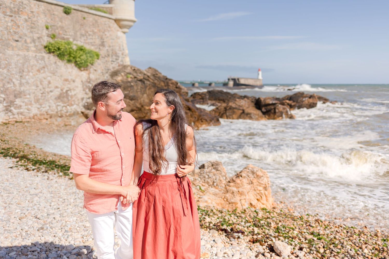 A couple shares a happy moment on a rocky beach with waves and a lighthouse in the distance.