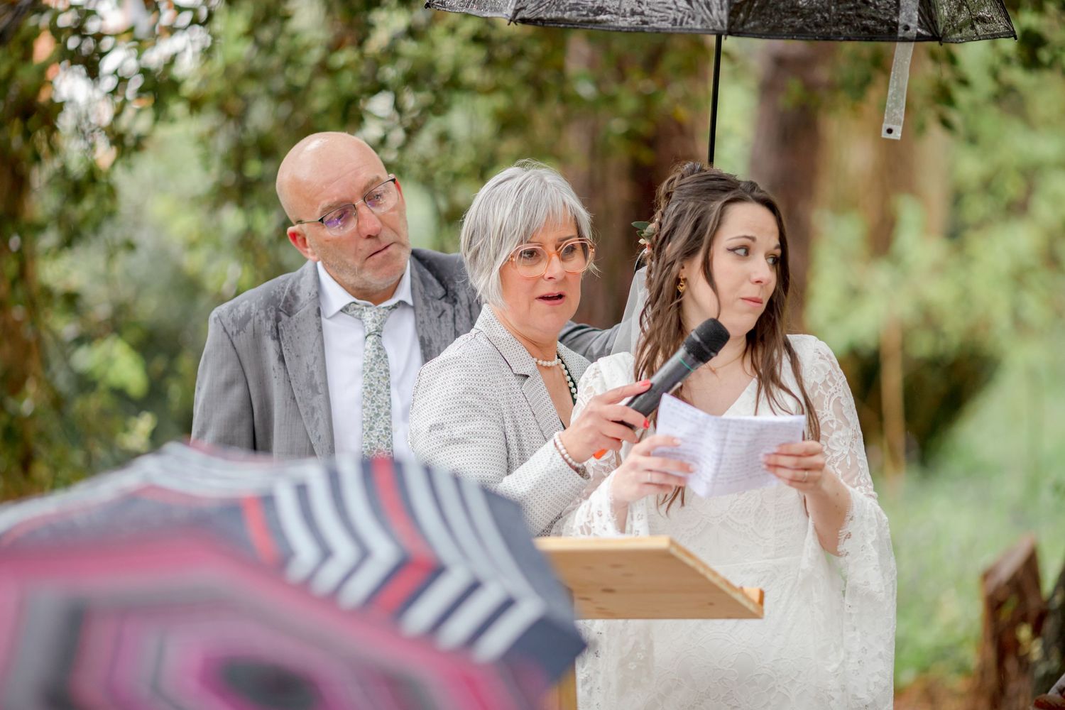 Wedding attendees sharing joyful moments under umbrellas during an outdoor forest ceremony.