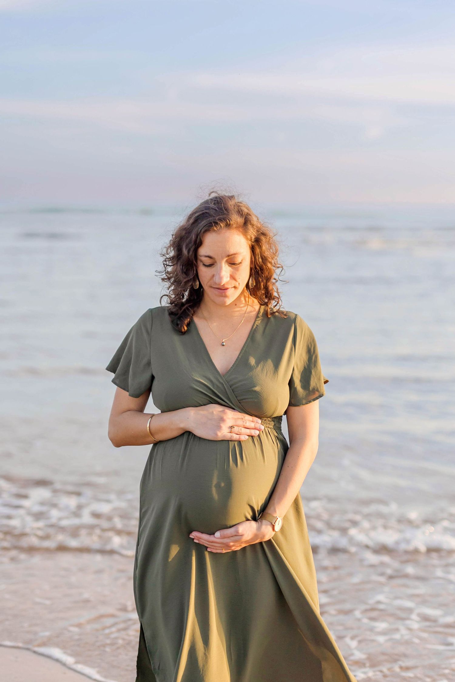 A person in a green wrap dress stands on the beach at sunset with gentle waves in the background.