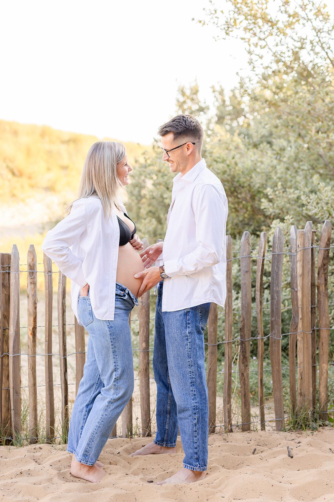 Couple in white shirts and blue jeans share tender moments on a sandy beach path with wooden fencing.
