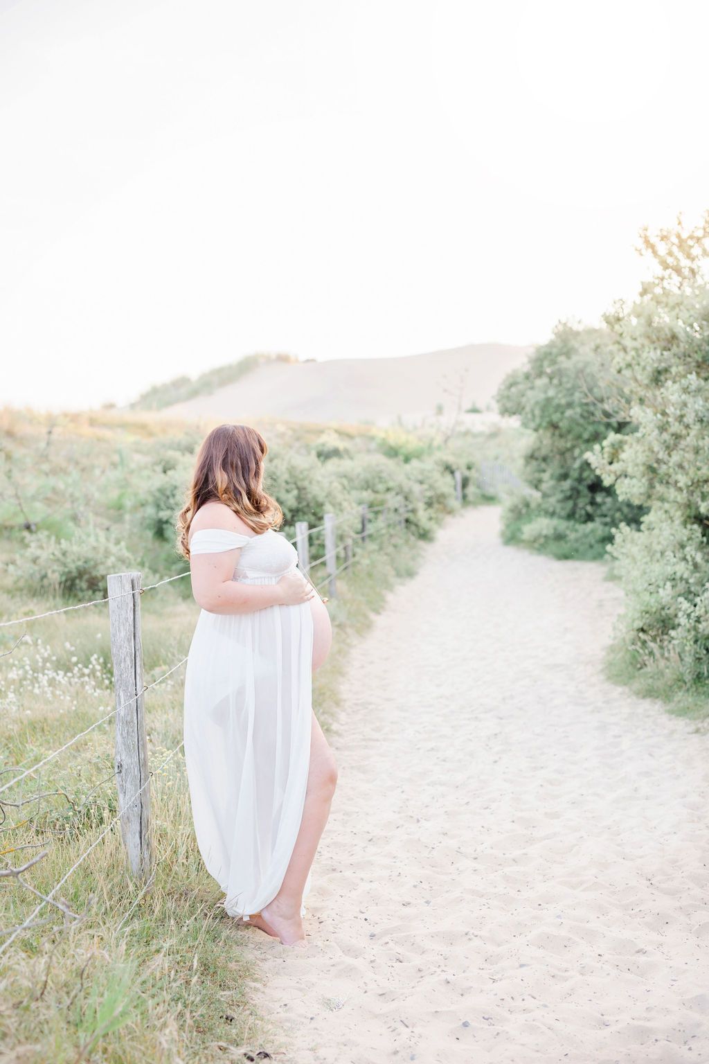 Ethereal beach maternity session on sandy path with coastal vegetation and wooden fence.