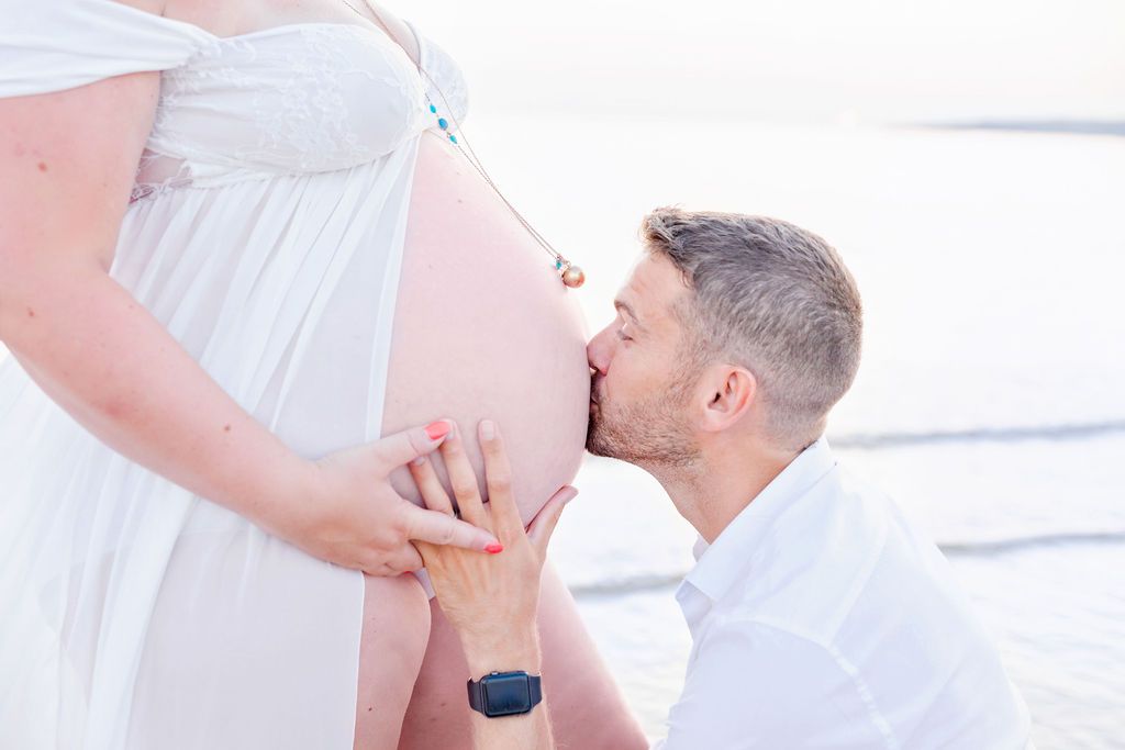 A romantic beach maternity photoshoot with a couple dressed in white and denim during sunset.