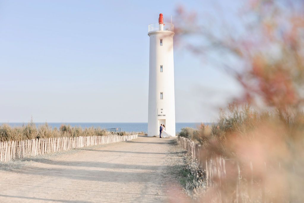 A white lighthouse tower stands along a coastal path with beach fencing and soft blurred foreground details.
