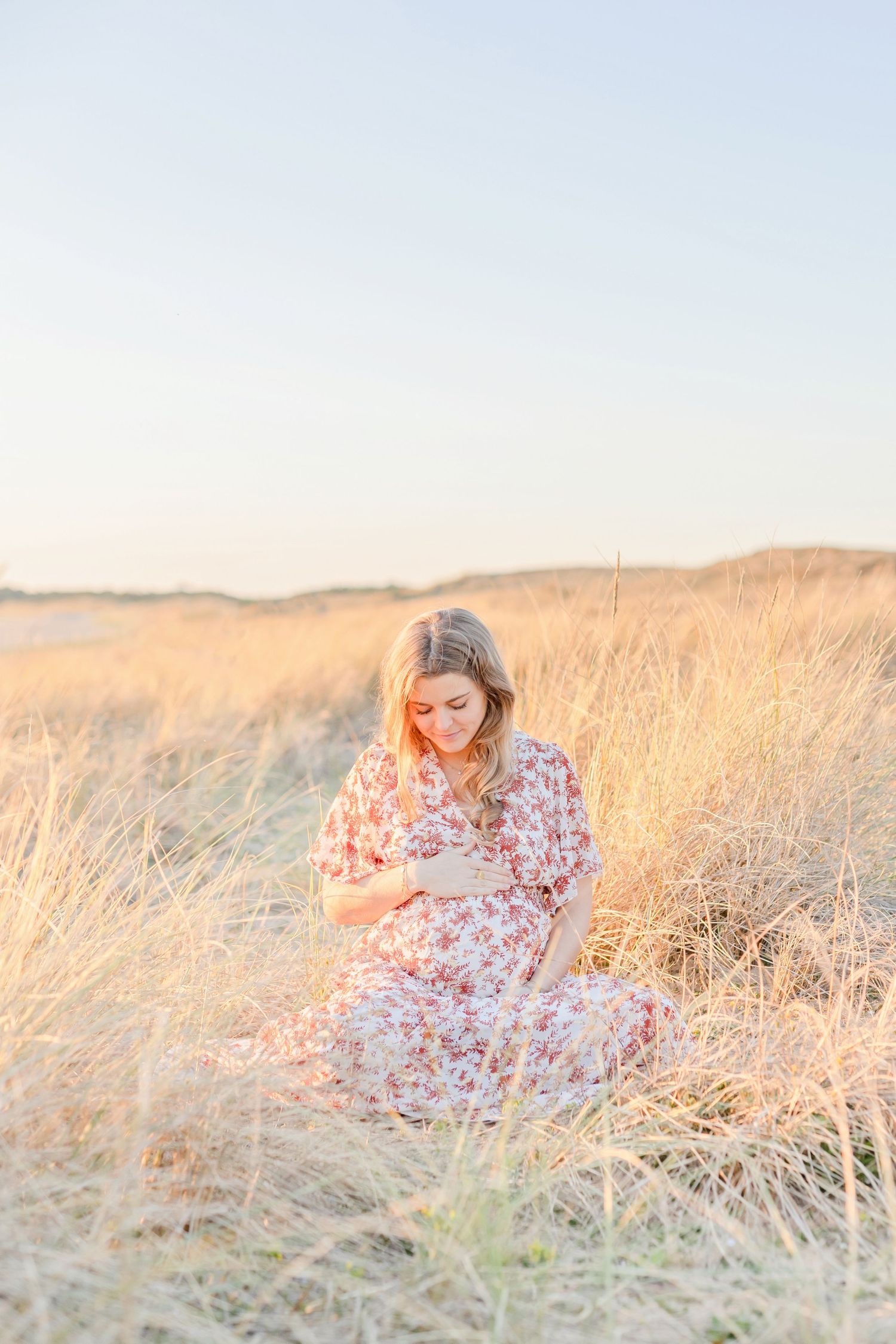 Woman in floral dress sitting peacefully in sunlit coastal grassland.