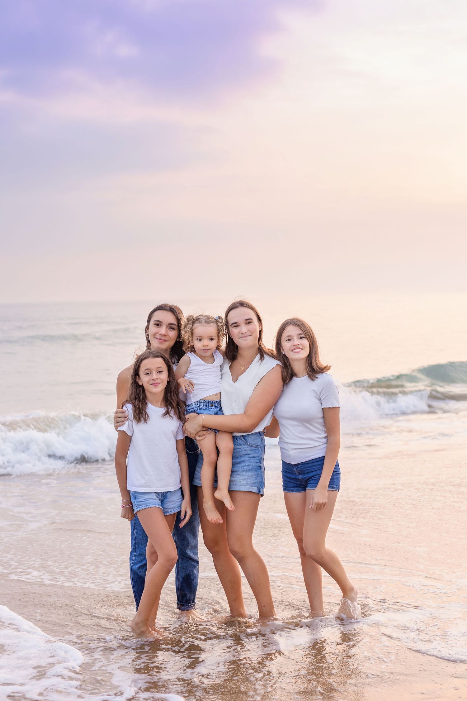 A group in matching white tops and denim enjoying a sunset moment at the beach with waves in the background.