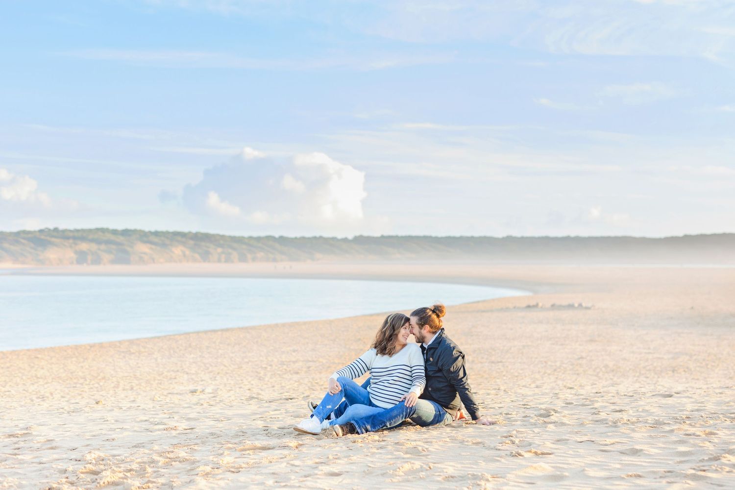 Couple sitting together on a sandy beach during golden hour with gentle waves in the background.