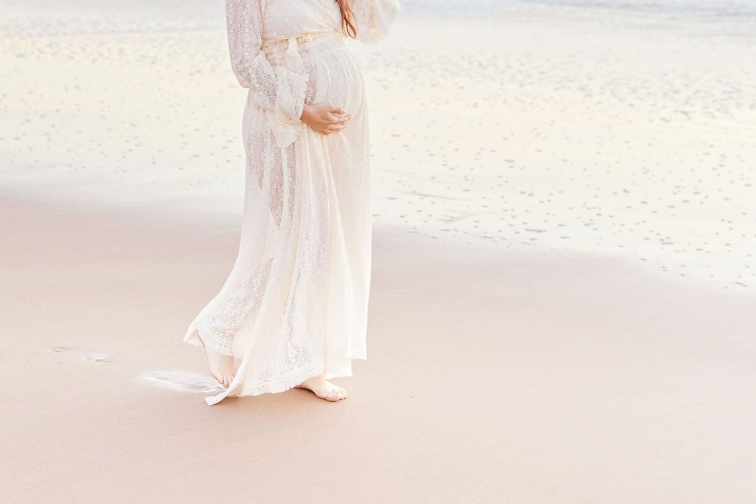 Ethereal silhouette of a pregnant woman in a flowing white dress on the beach.