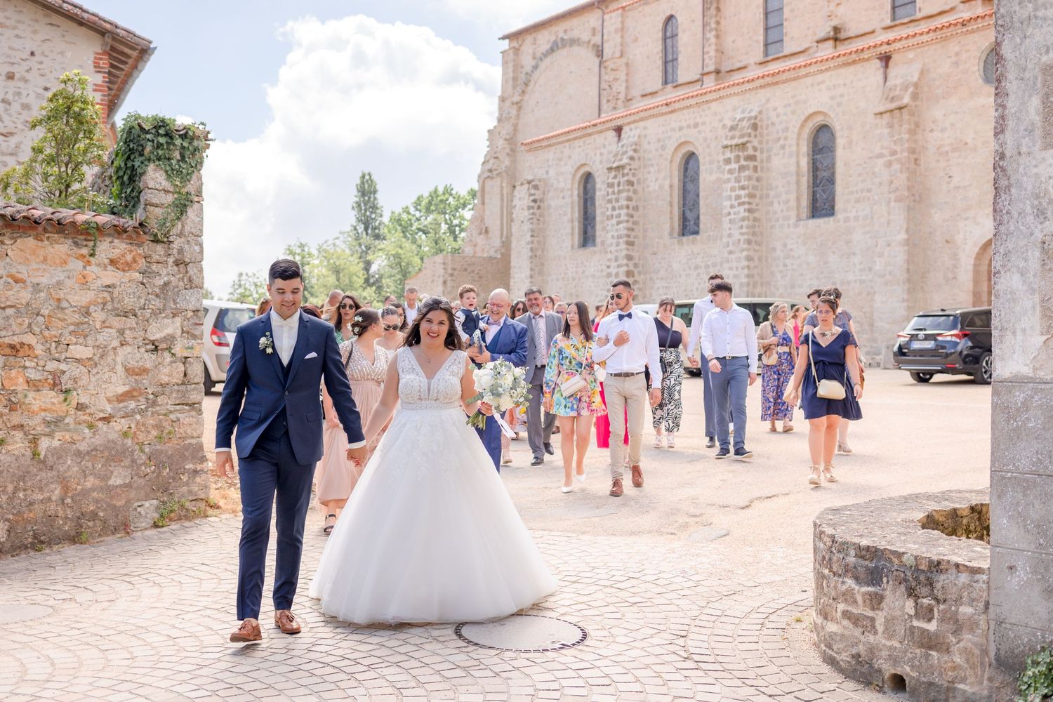 Couple walks away from stone church building followed by wedding guests on cobblestone path.