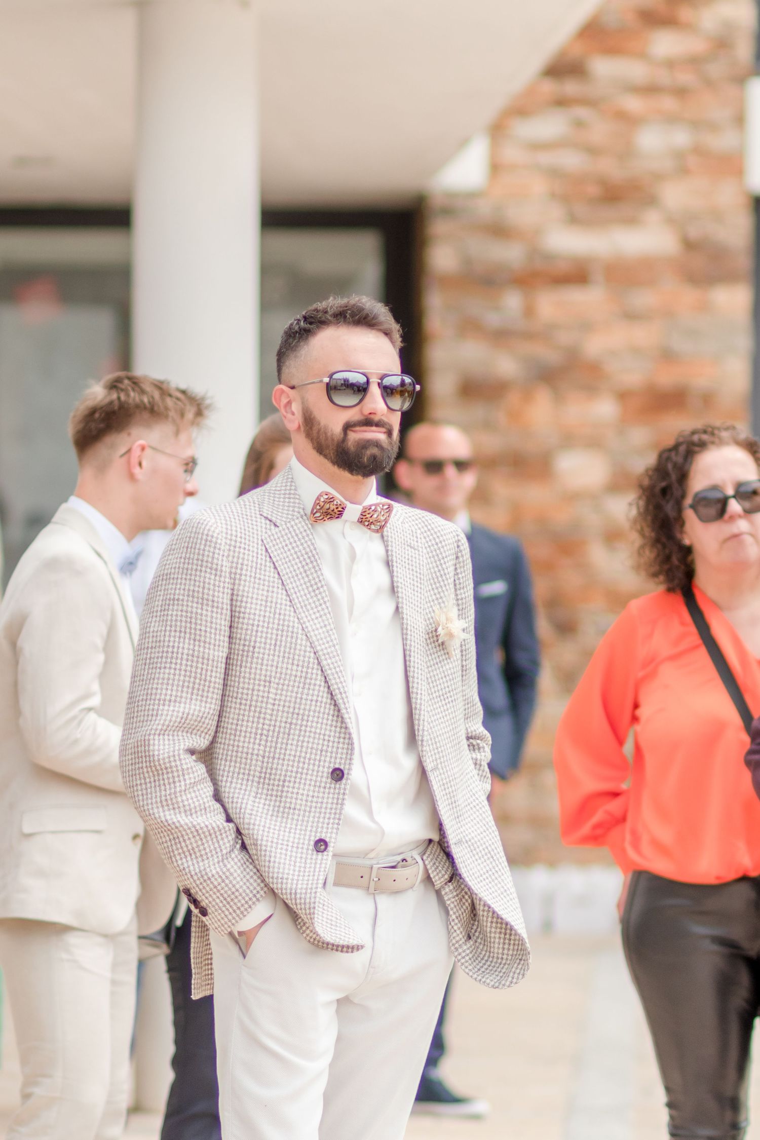 Wedding party members in white and checkered suits stand together outside a brick building.