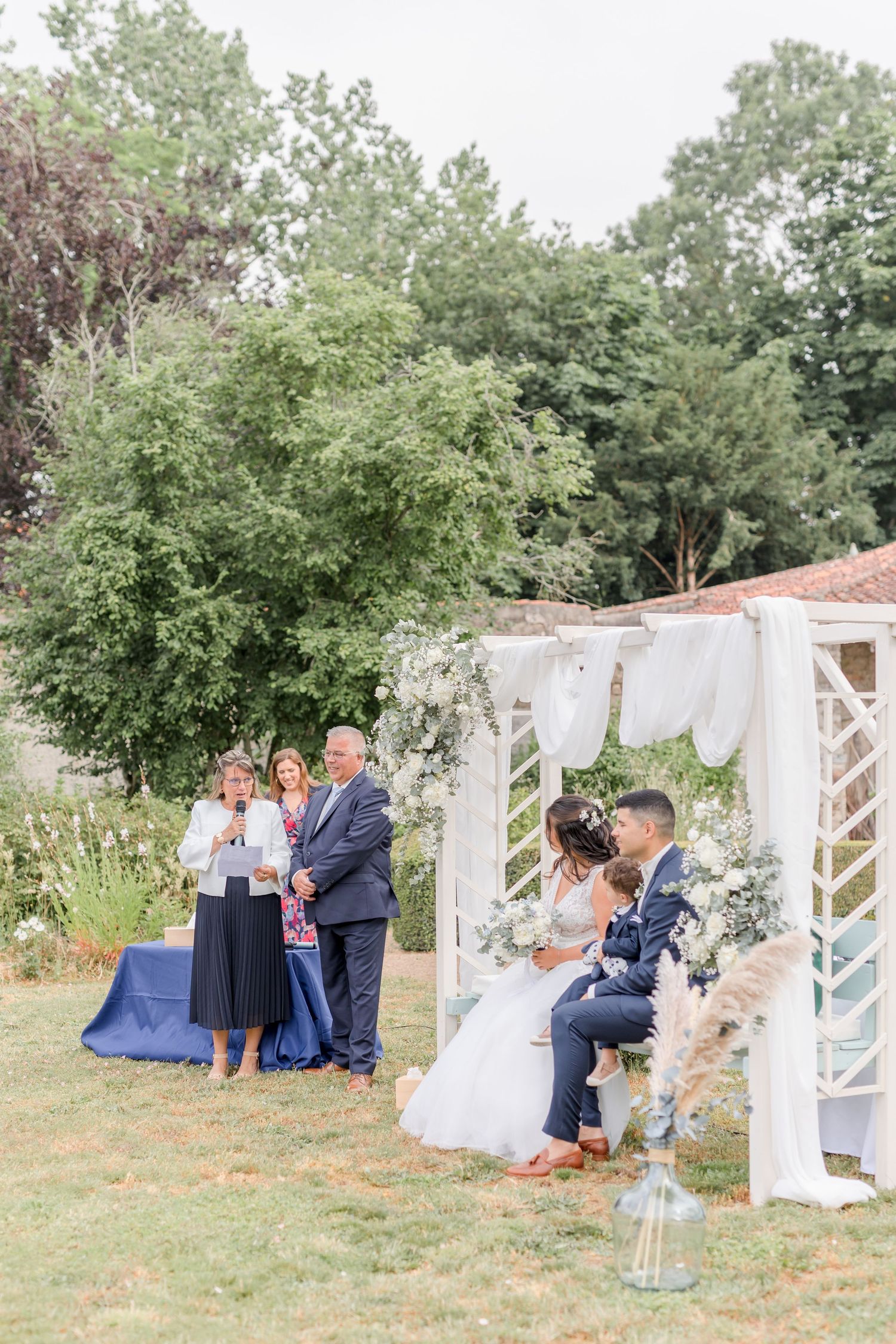 Wedding ceremony scene with decorated white arch and guests seated on lawn.