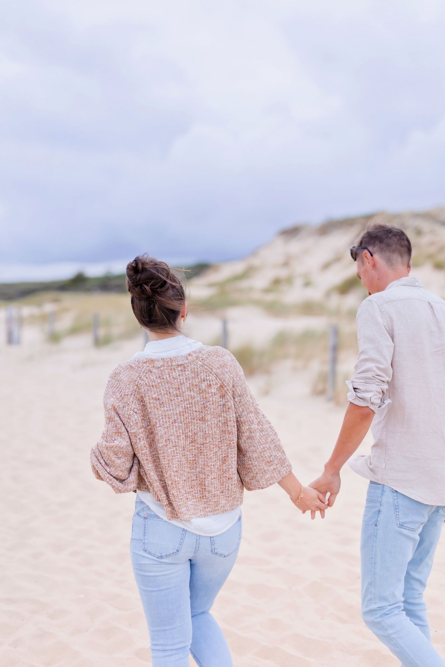 Couple walking hand in hand along sandy beach with dunes in the background.