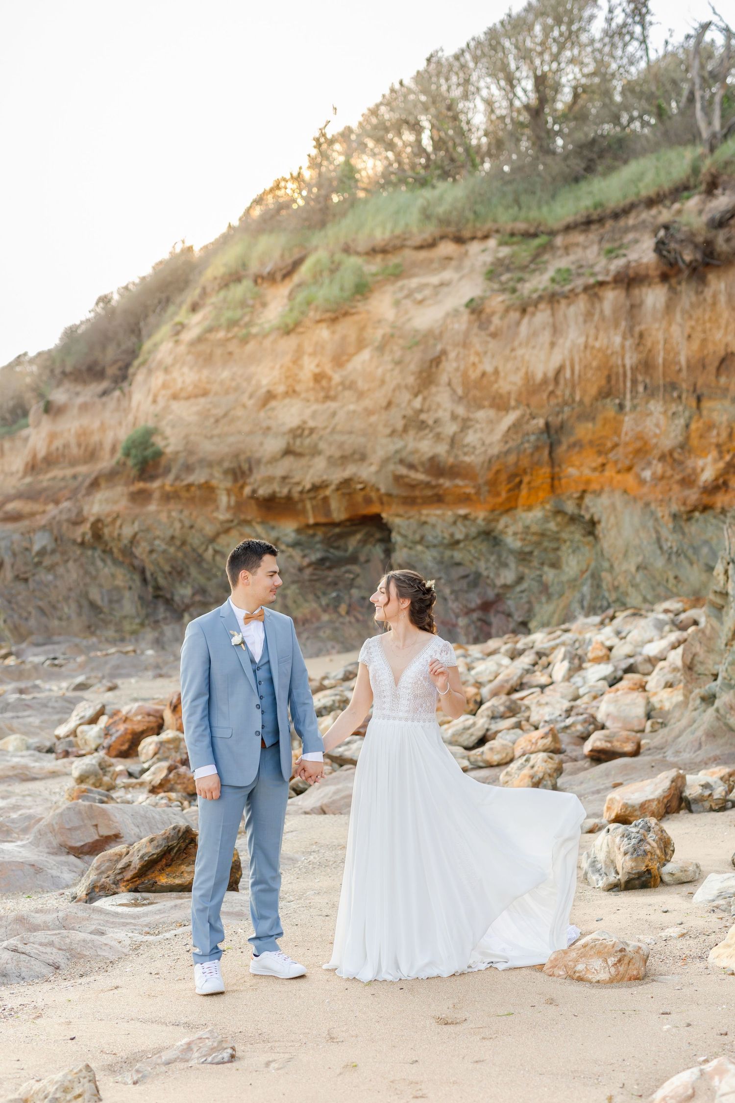 A couple in wedding attire sits on rocky beach terrain near coastal cliffs.