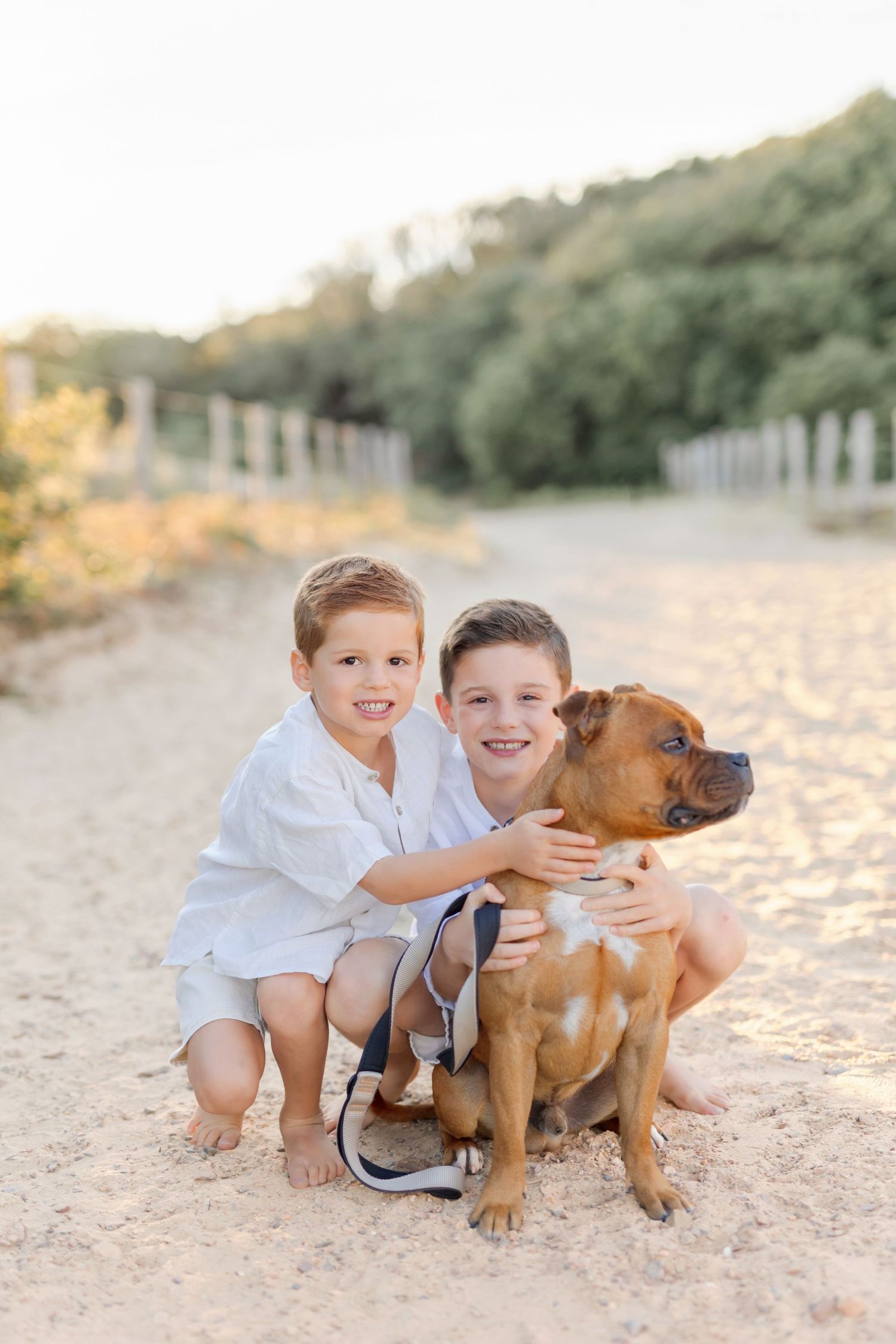 Two children crouching next to brown puppy on beach sand.