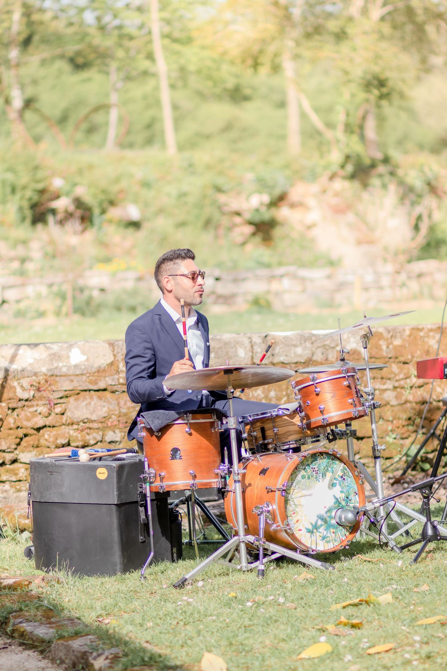 Musicians playing drums and instruments set up outdoors against a stone wall in natural sunlight.