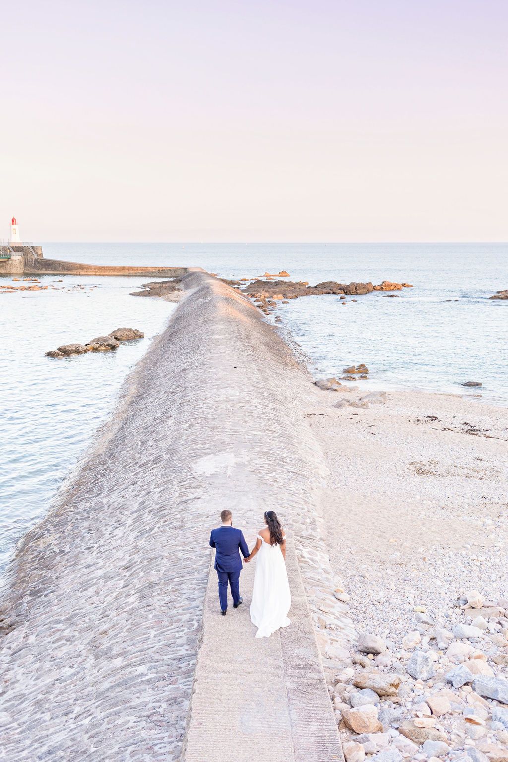 Couple in wedding attire walk hand in hand along a rocky coastal path by the ocean.