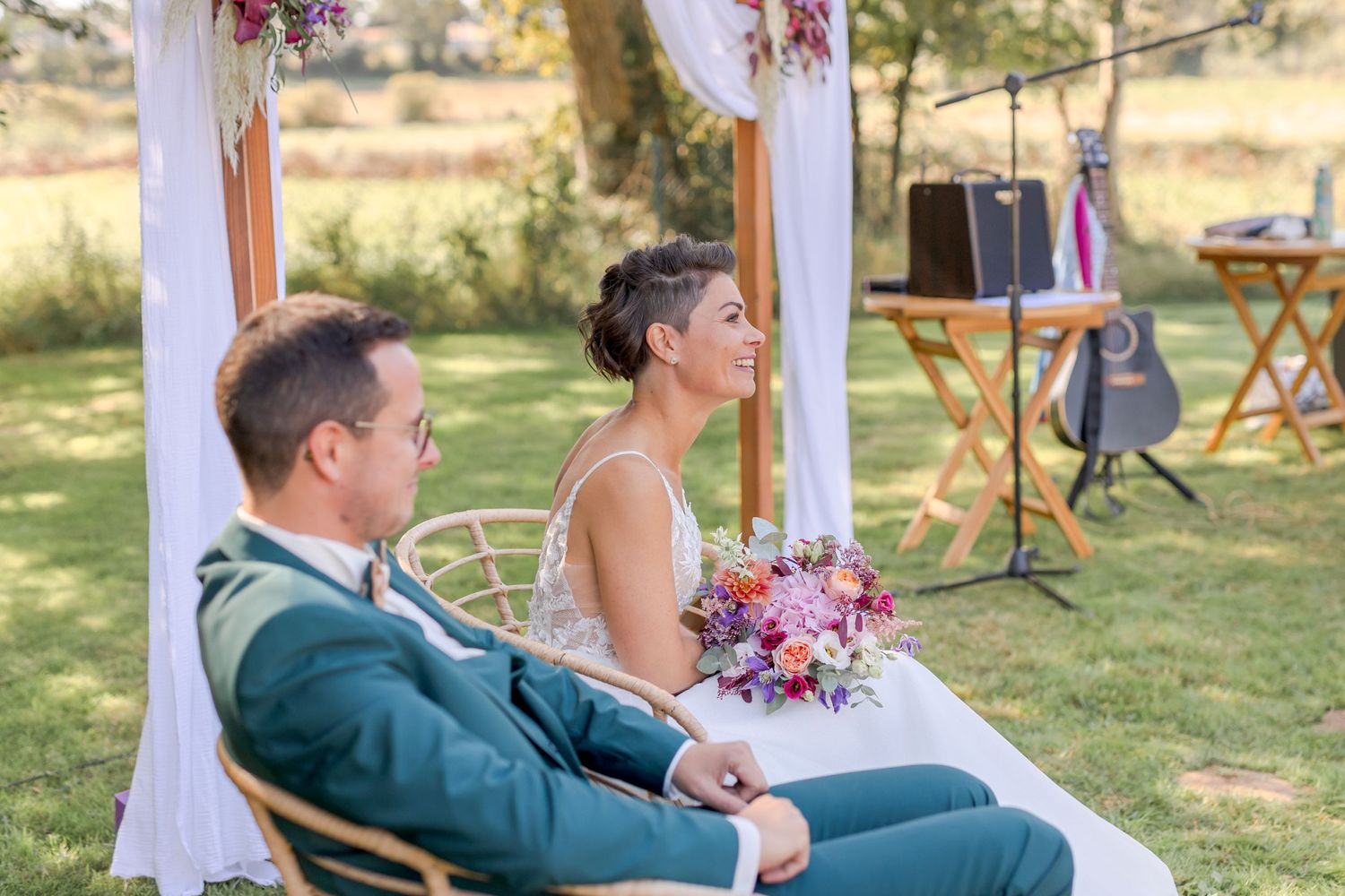 Couple seated under floral arch during outdoor wedding ceremony.