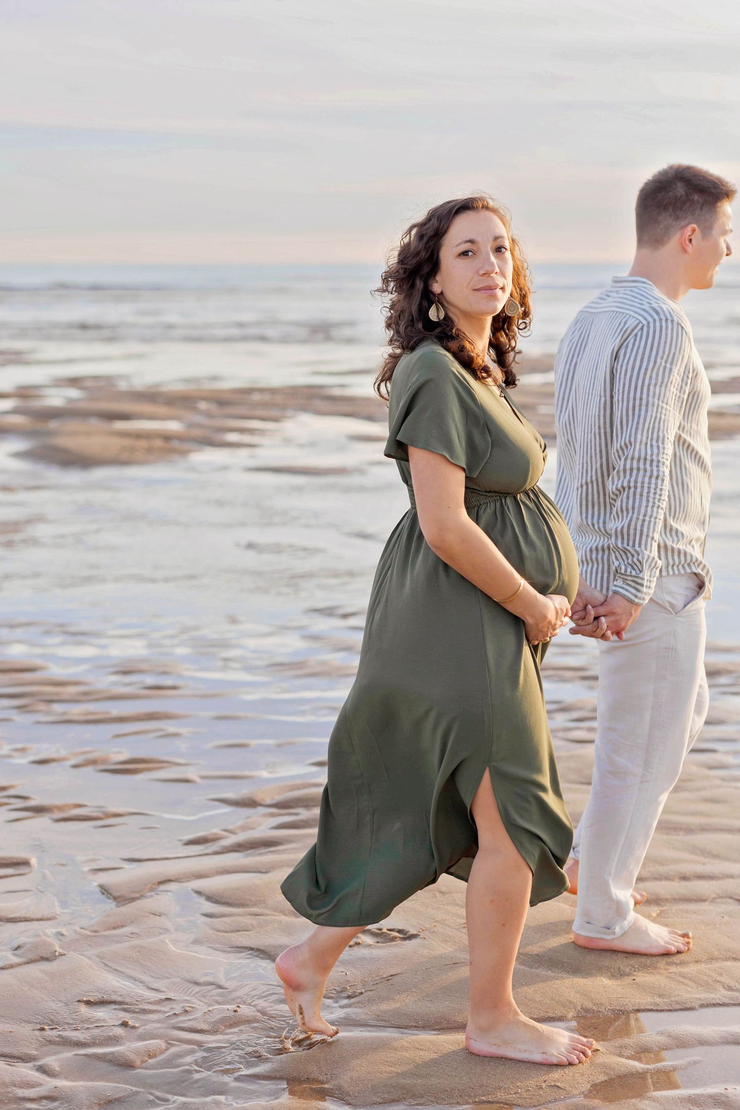 A person in a flowing green dress stands barefoot on the beach while another looks towards the ocean.