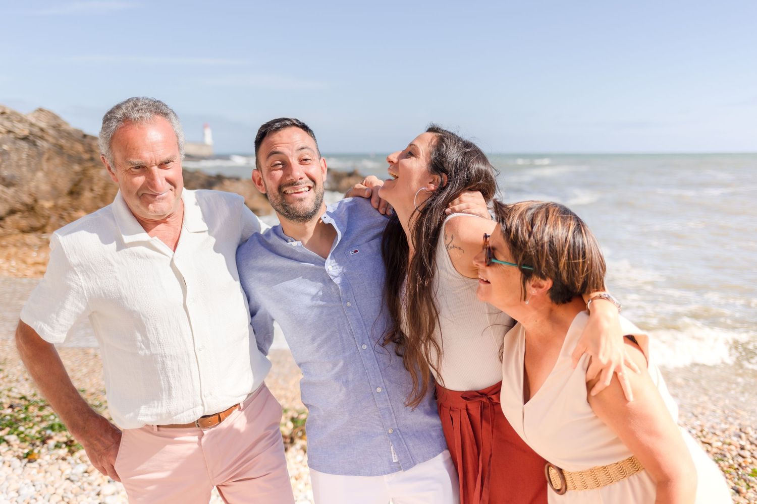 Four people laugh together on a sunny beach with waves and rocky coastline behind them.
