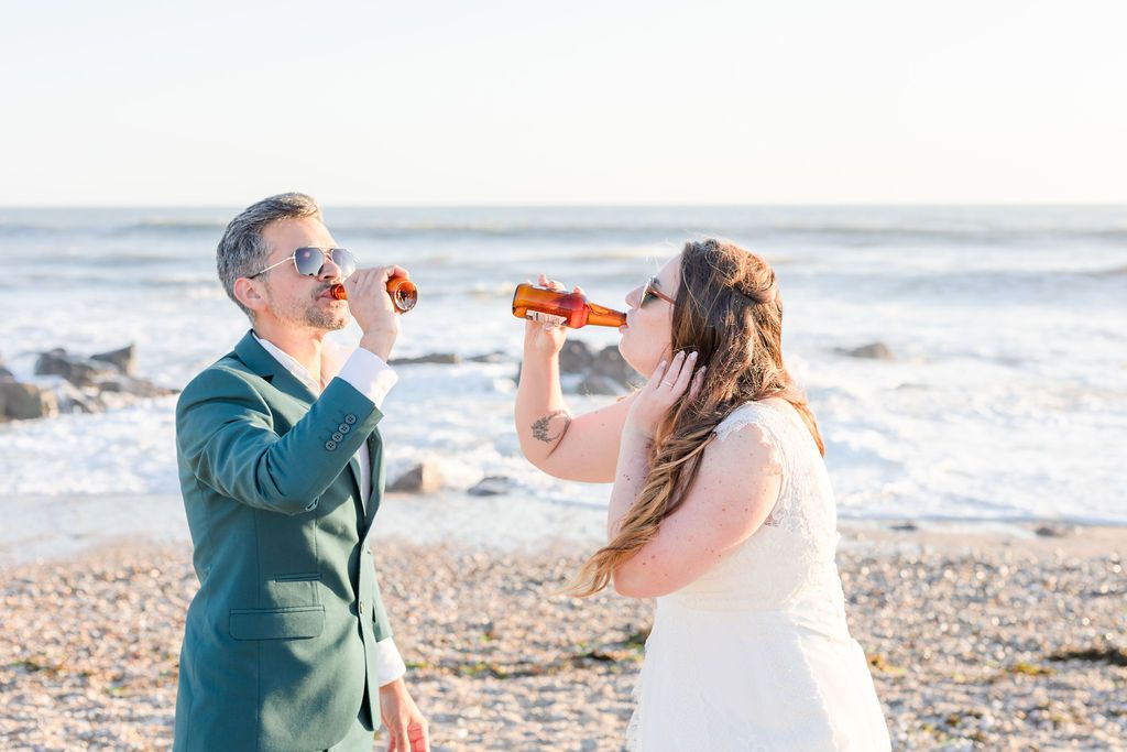 Wedding couple playfully drinking beer together on a sunny beach during their celebration.