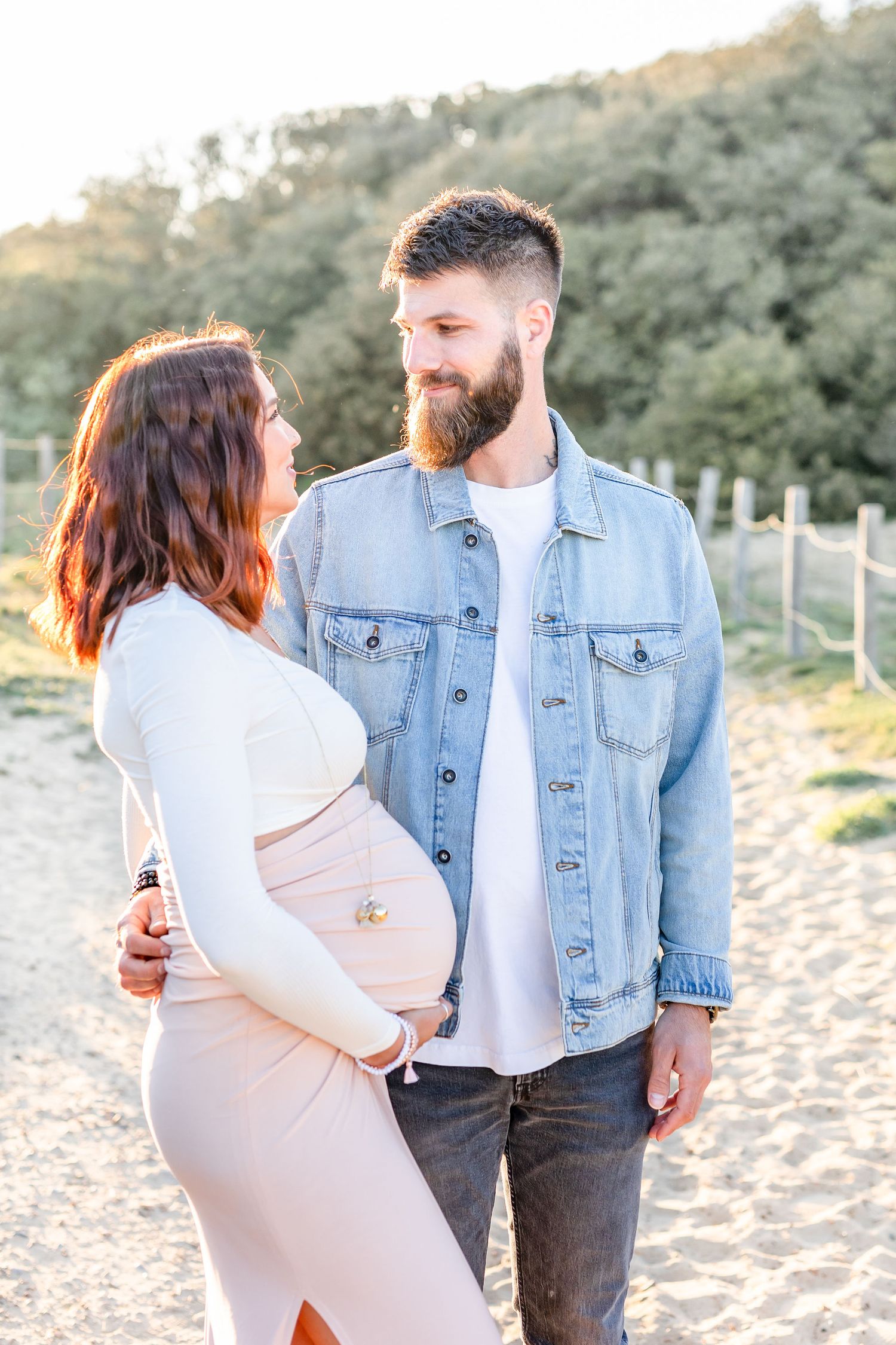 Couple in denim and white embrace on sandy beach during maternity photoshoot.