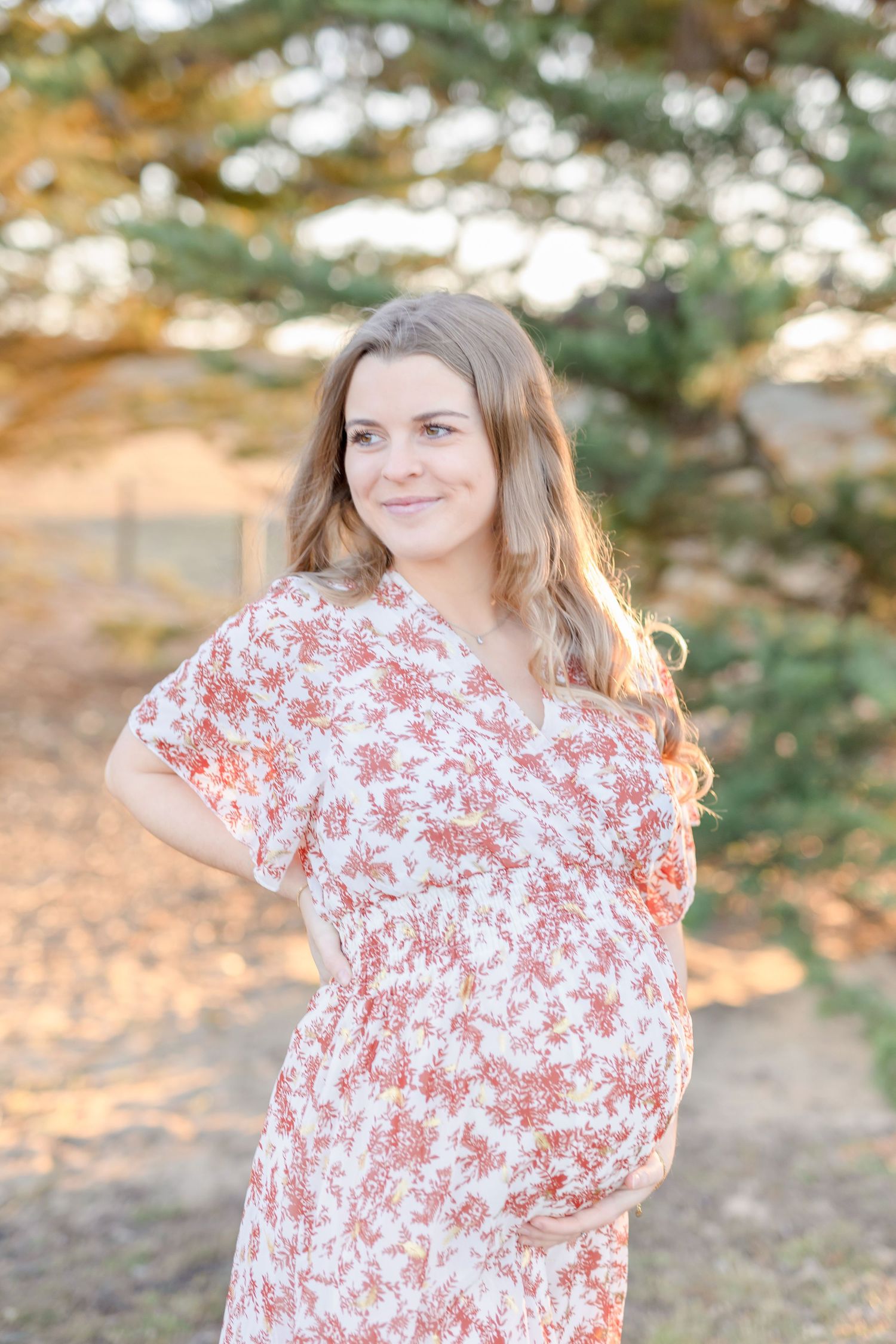 Expectant woman in red and white floral dress smiling outdoors during golden hour.