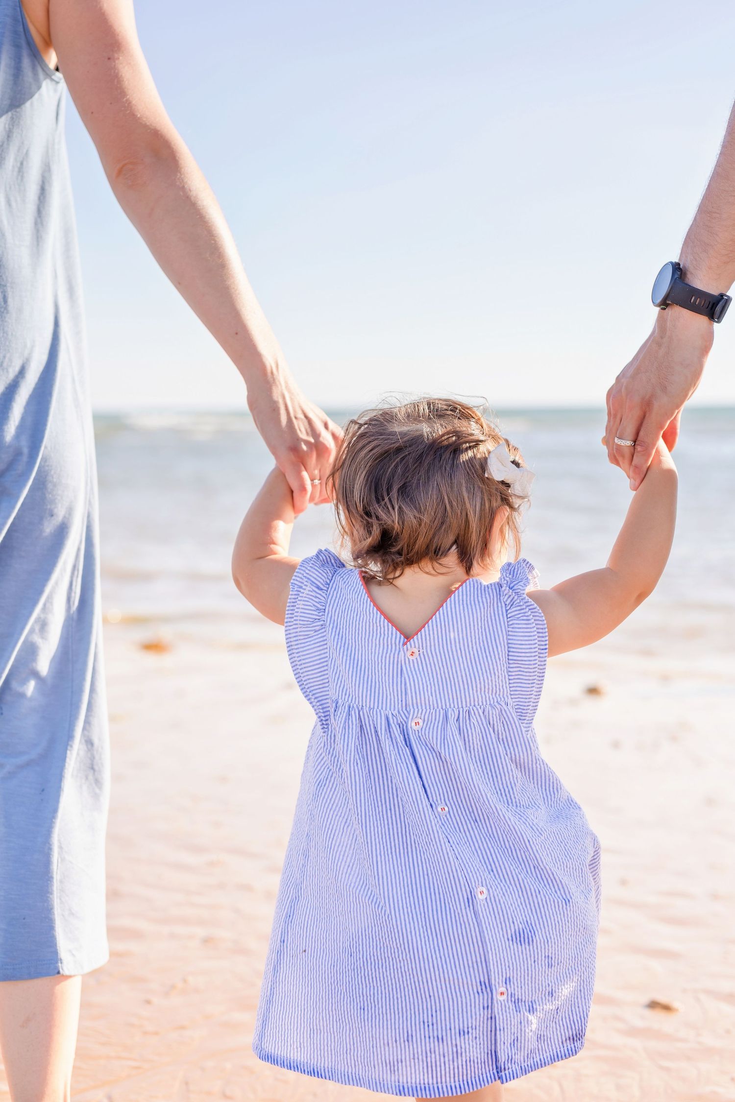 Small child in striped dress holding hands with adults while walking on beach.