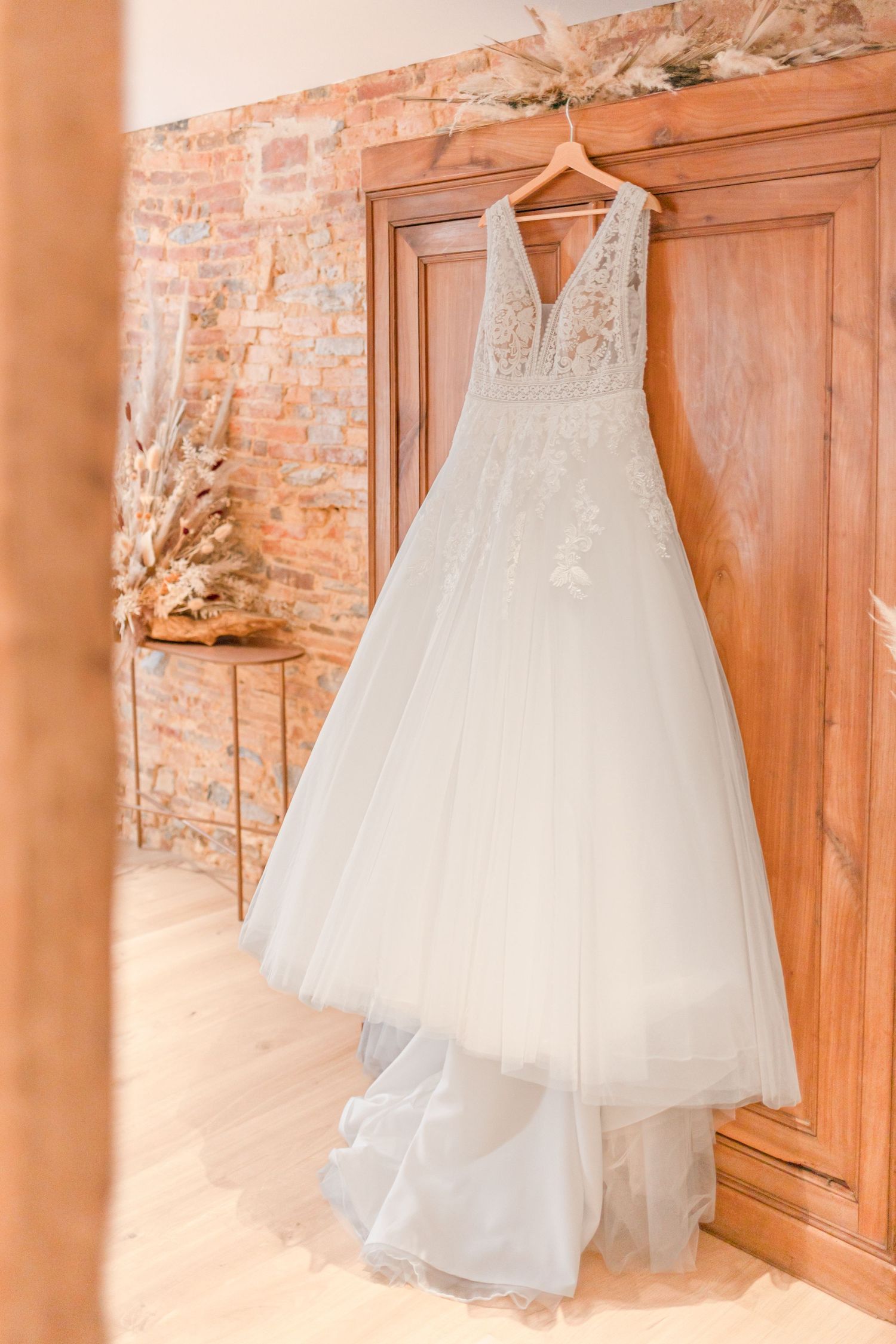 Elegant white tulle wedding dress with lace details hanging on wooden door against exposed brick wall.