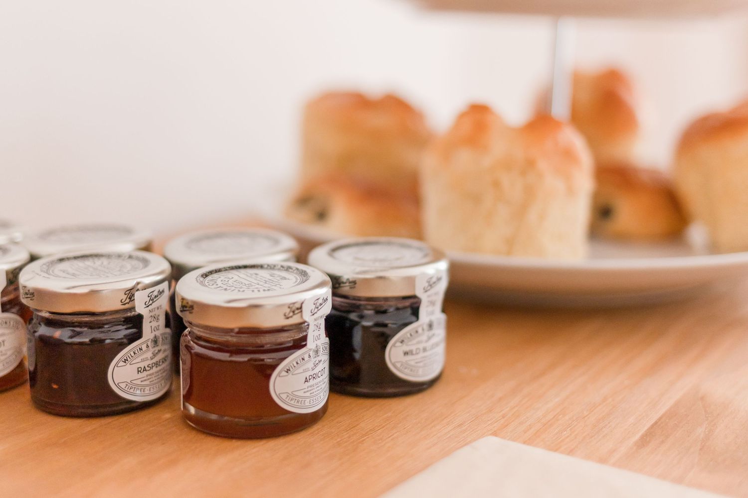 Three small jars of preserves or jam lined up on a wooden table with pastries in the background.