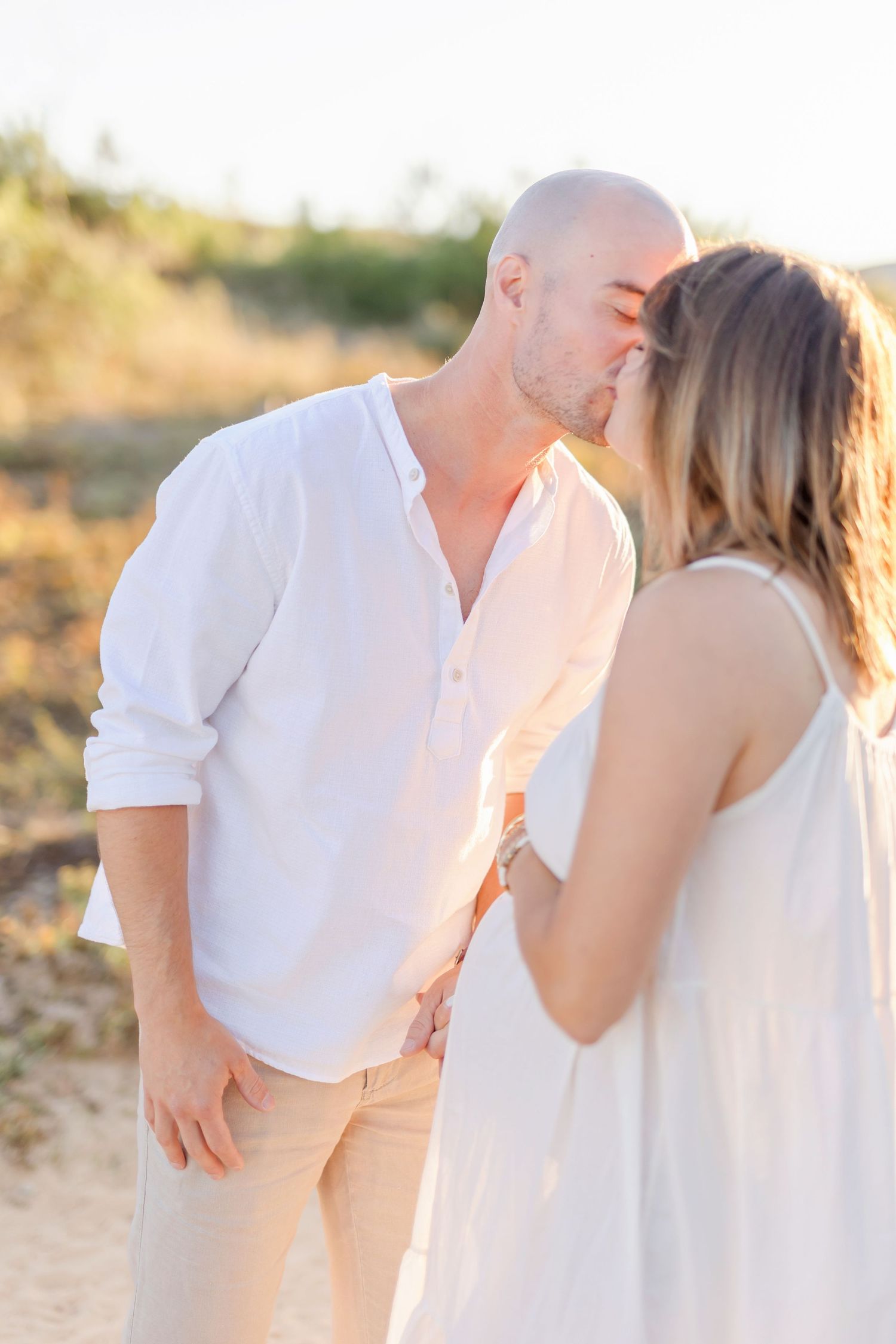 Candid maternity moment between couple wearing white and beige on dirt path at sunset.