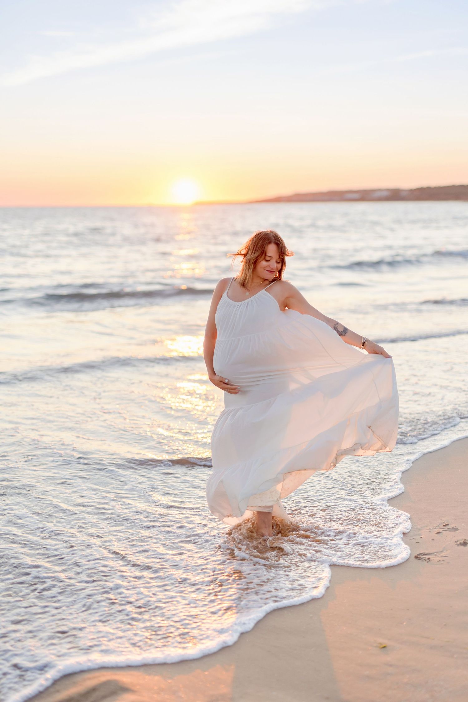 Woman in flowing white dress twirls on beach at sunset with waves lapping at shore.