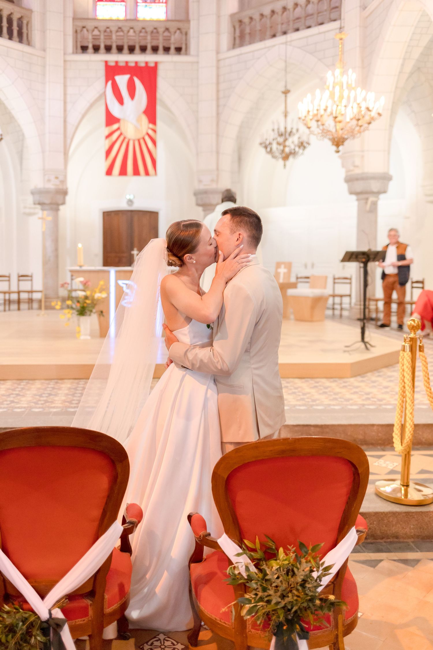 A romantic wedding kiss in a grand church with red banners and elegant red chairs.
