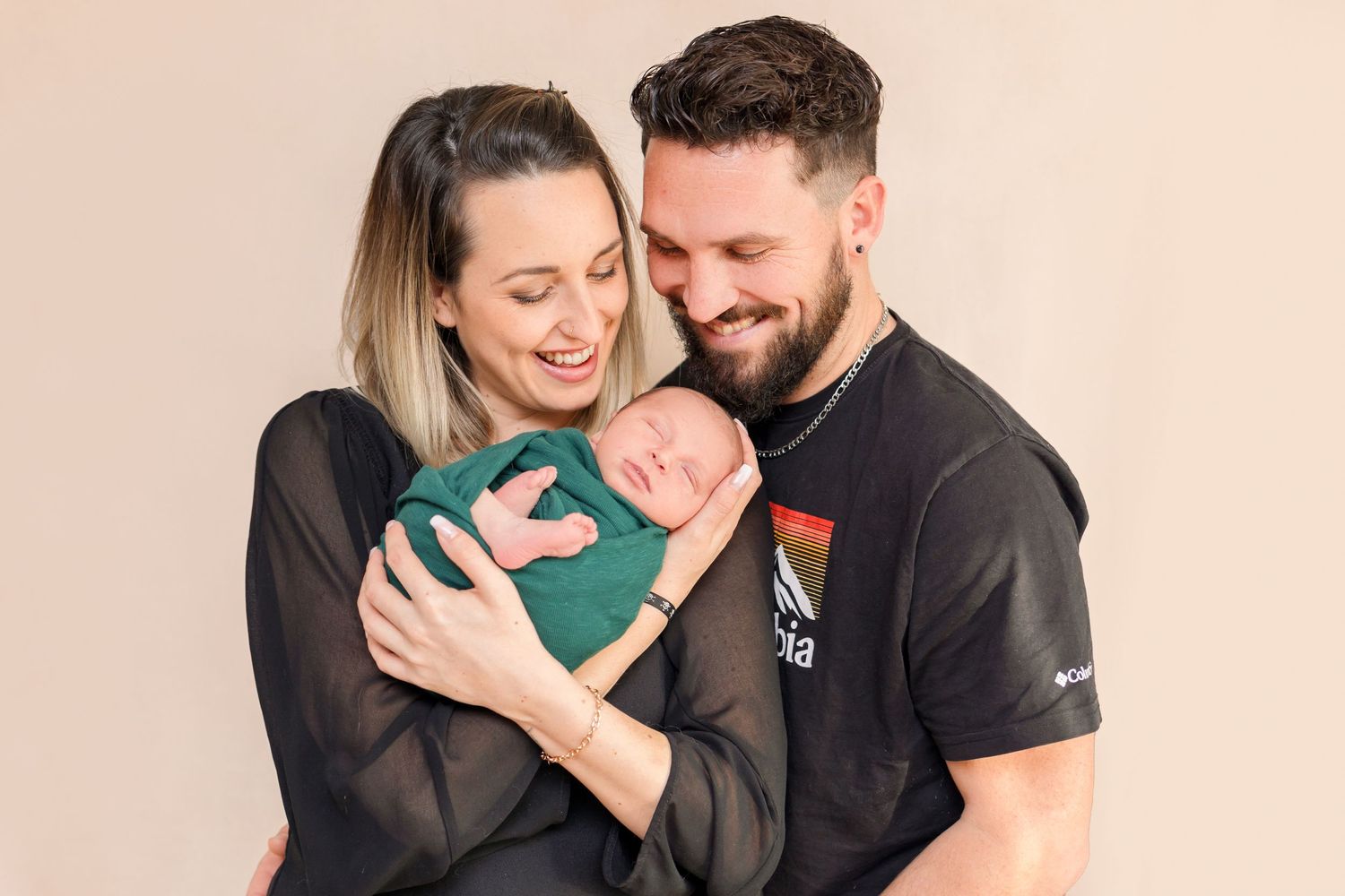 Joyful family moment with sleeping newborn wrapped in green against neutral backdrop.