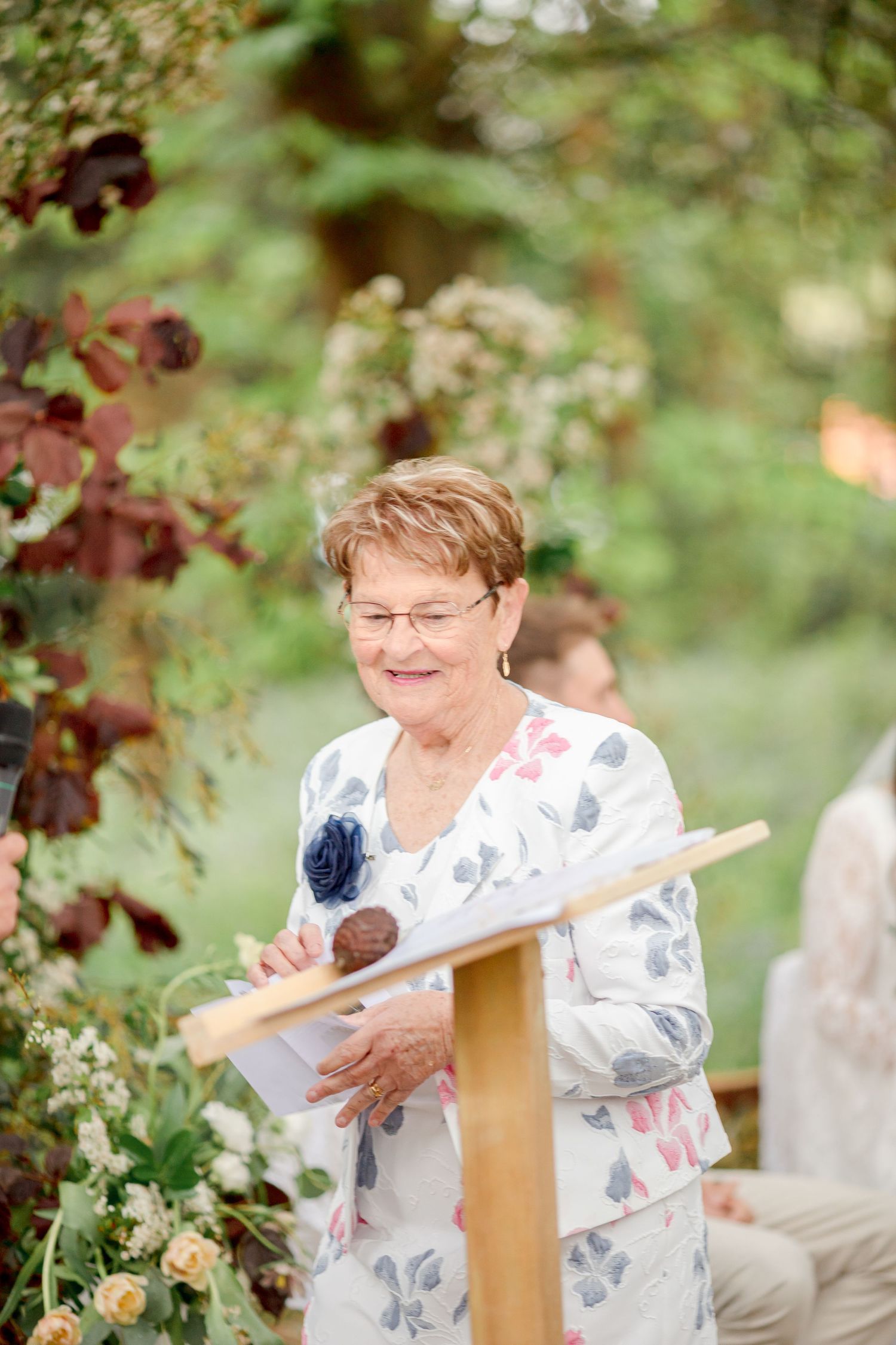 Speaker stands at wooden podium during outdoor ceremony surrounded by greenery.