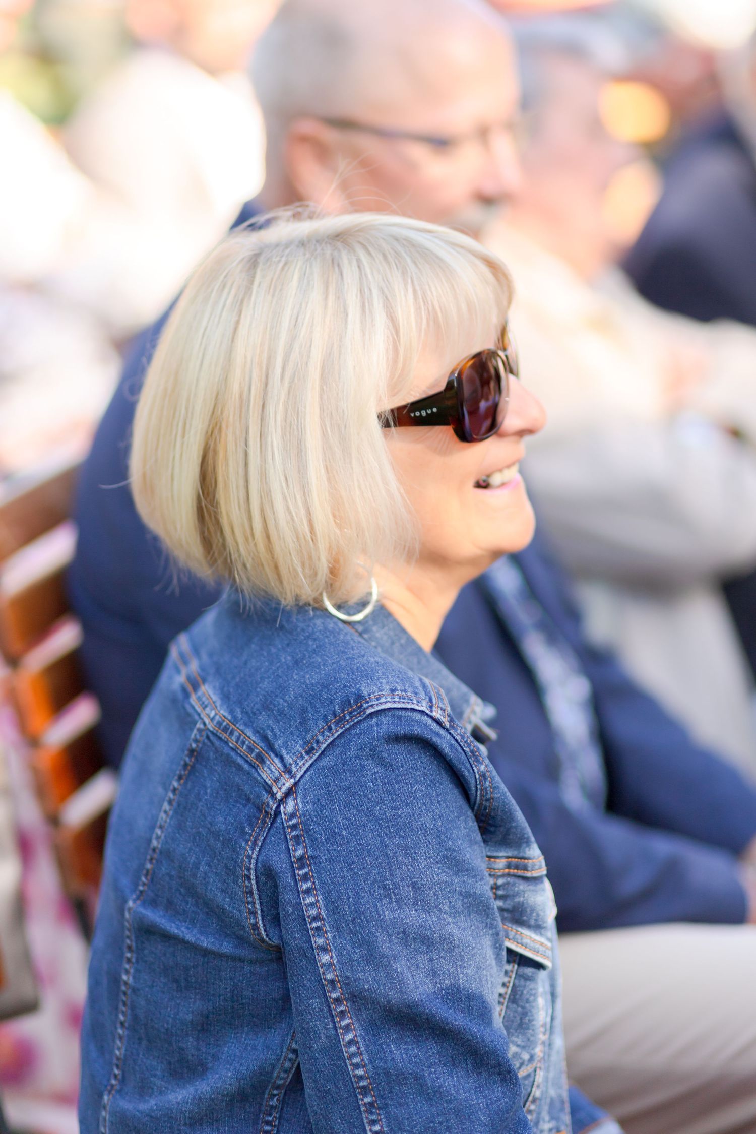 Wedding guest in denim jacket and sunglasses smiles while enjoying outdoor ceremony.