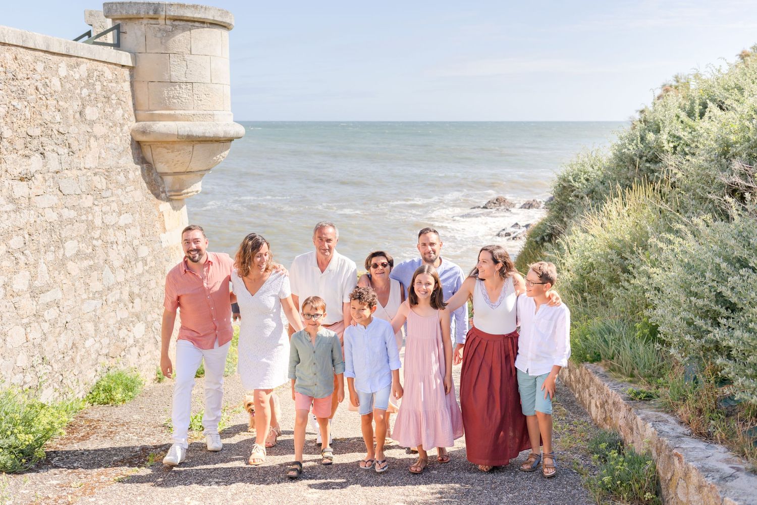A group poses together on a coastal path near a stone fortress wall overlooking the ocean.