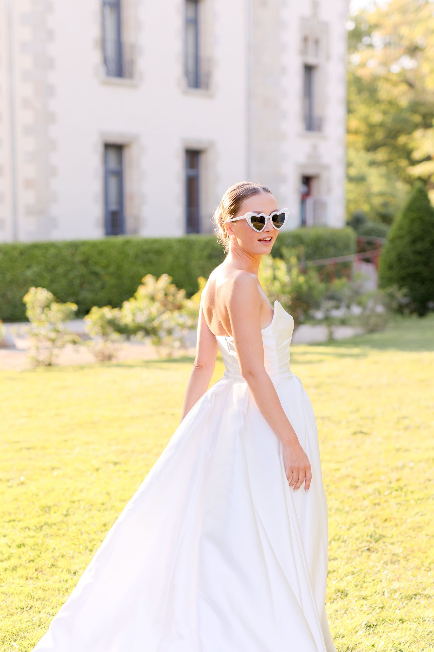 Bride in sunglasses twirls in elegant white dress on sunny castle grounds.