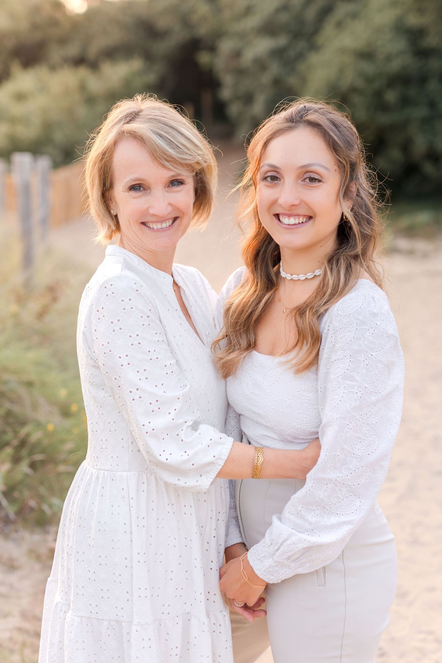 Two women in white dresses smiling and embracing on outdoor path during sunset.
