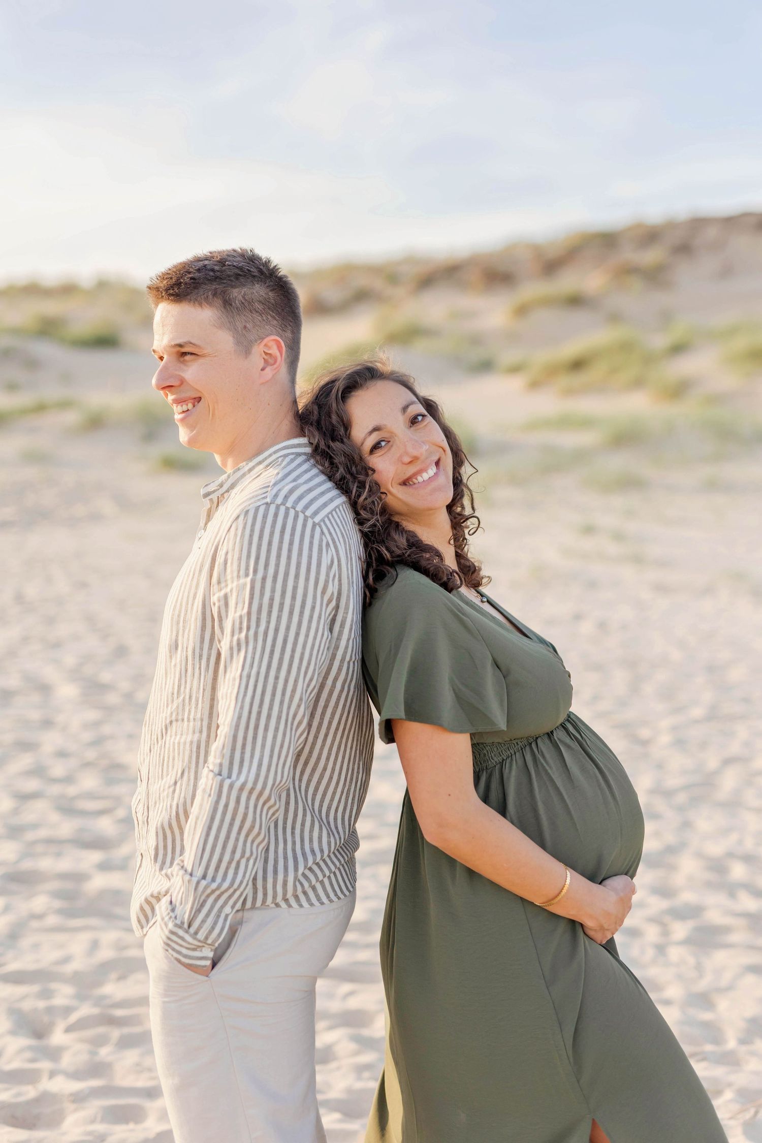 A couple stands together on beach during sunset maternity photoshoot.