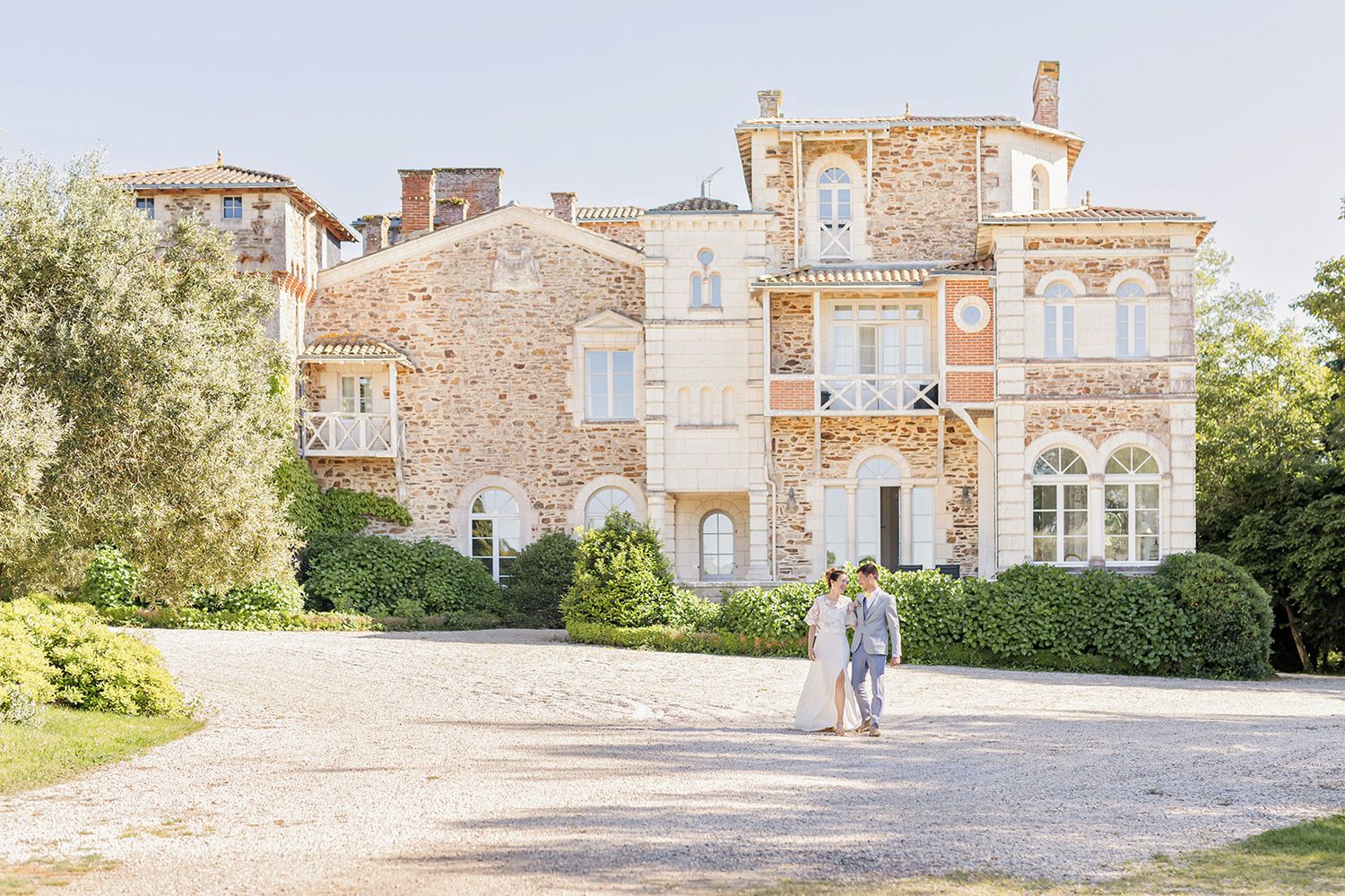 Elegant Victorian mansion with stone facade and ornate details stands on a gravel path surrounded by lush gardens.