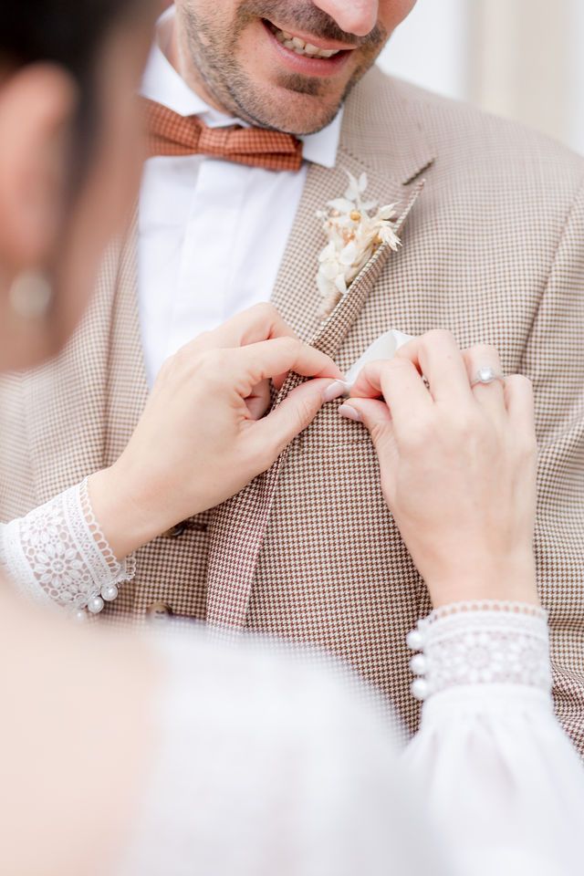 Hands with lace sleeves adjust a coral bowtie on beige suit with floral boutonniere.