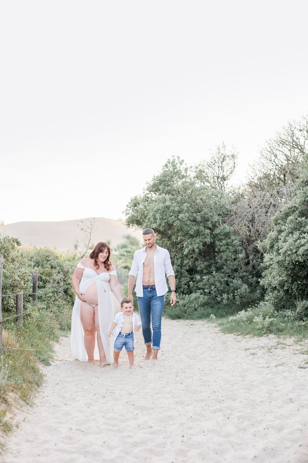 Family walking together on a sandy beach path surrounded by coastal vegetation on a bright day.