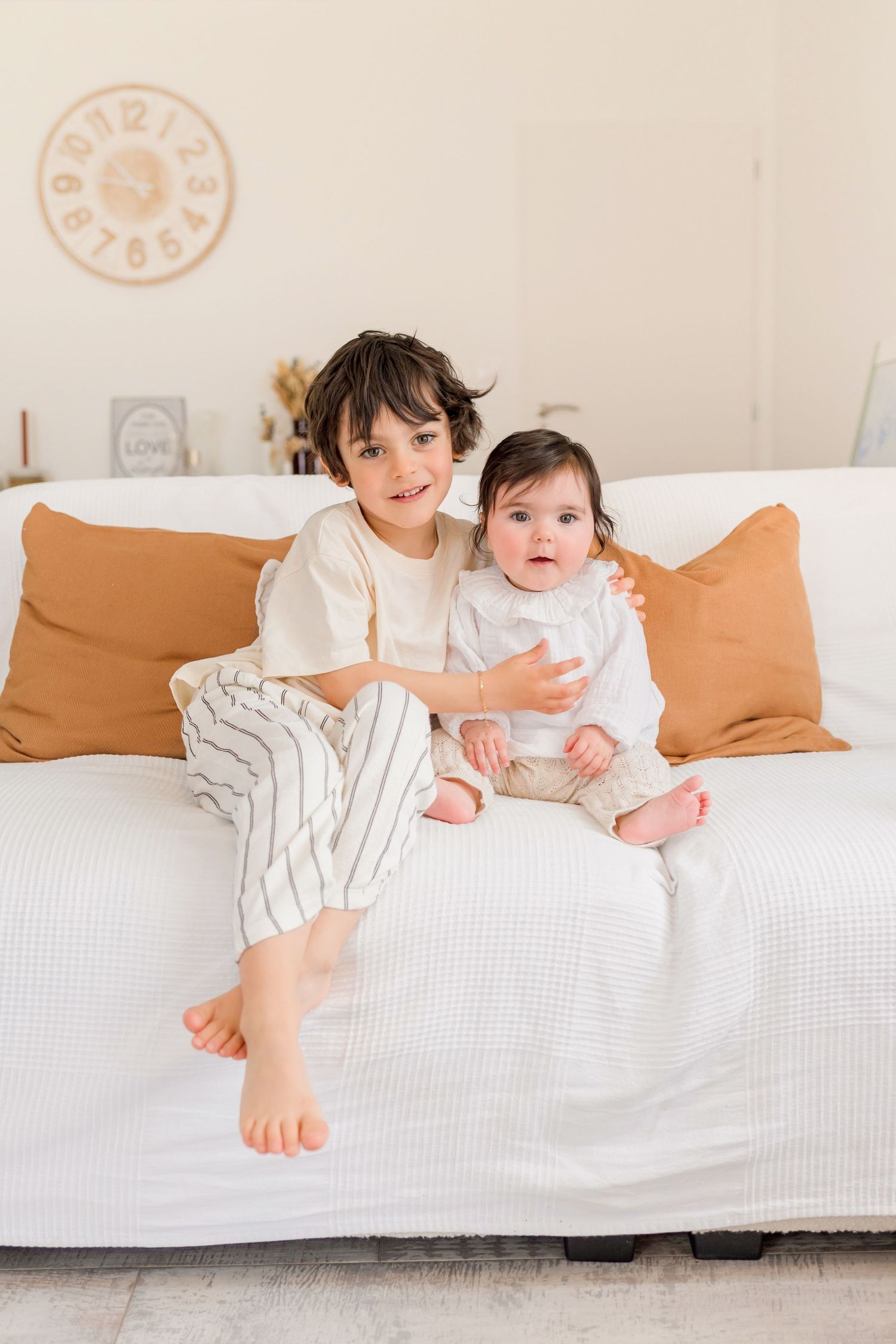 Two children sitting together on a white couch with orange throw pillows in a bright room.