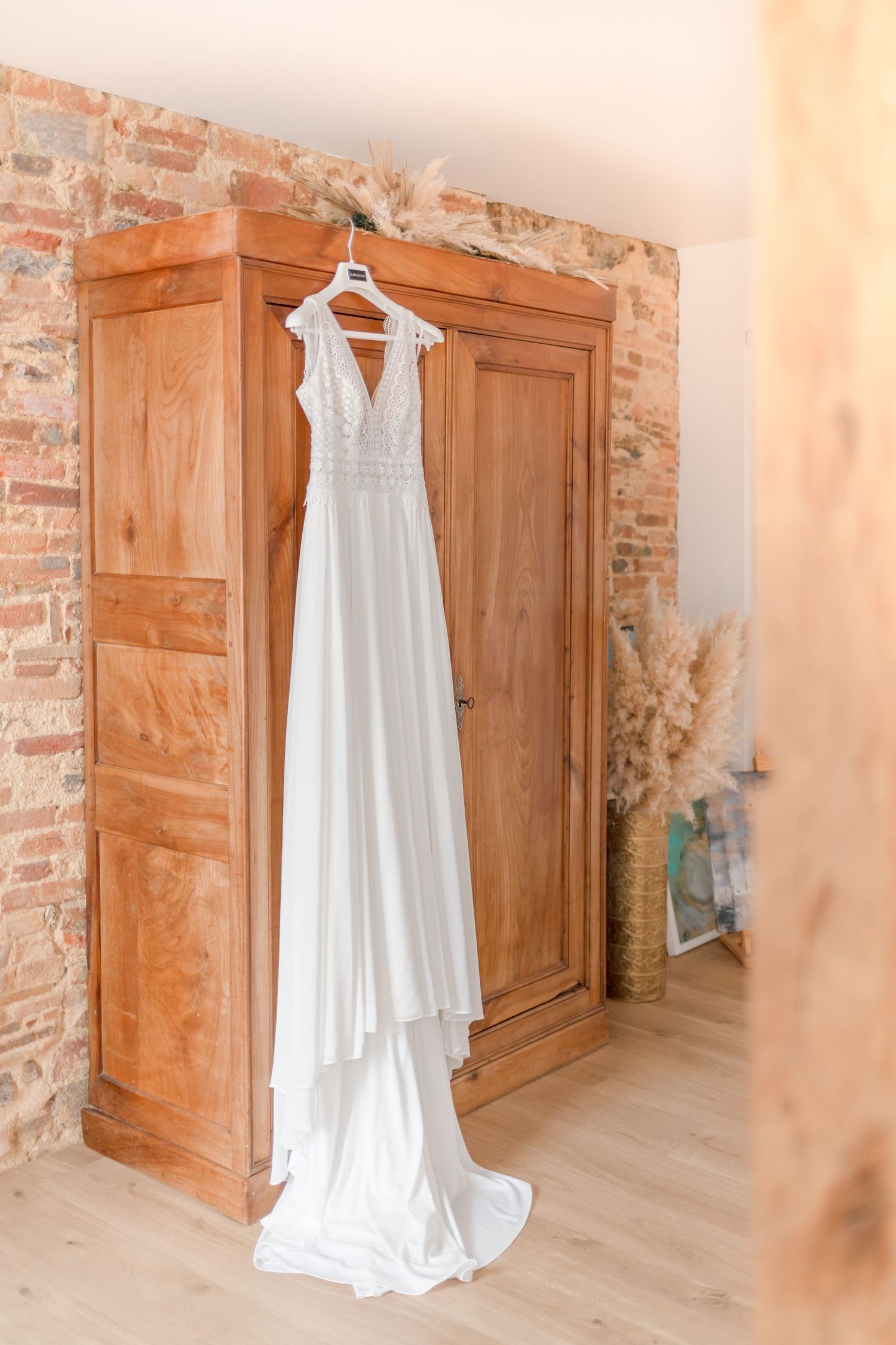 Full-length white wedding gown displayed against wooden armoire with exposed brick wall.