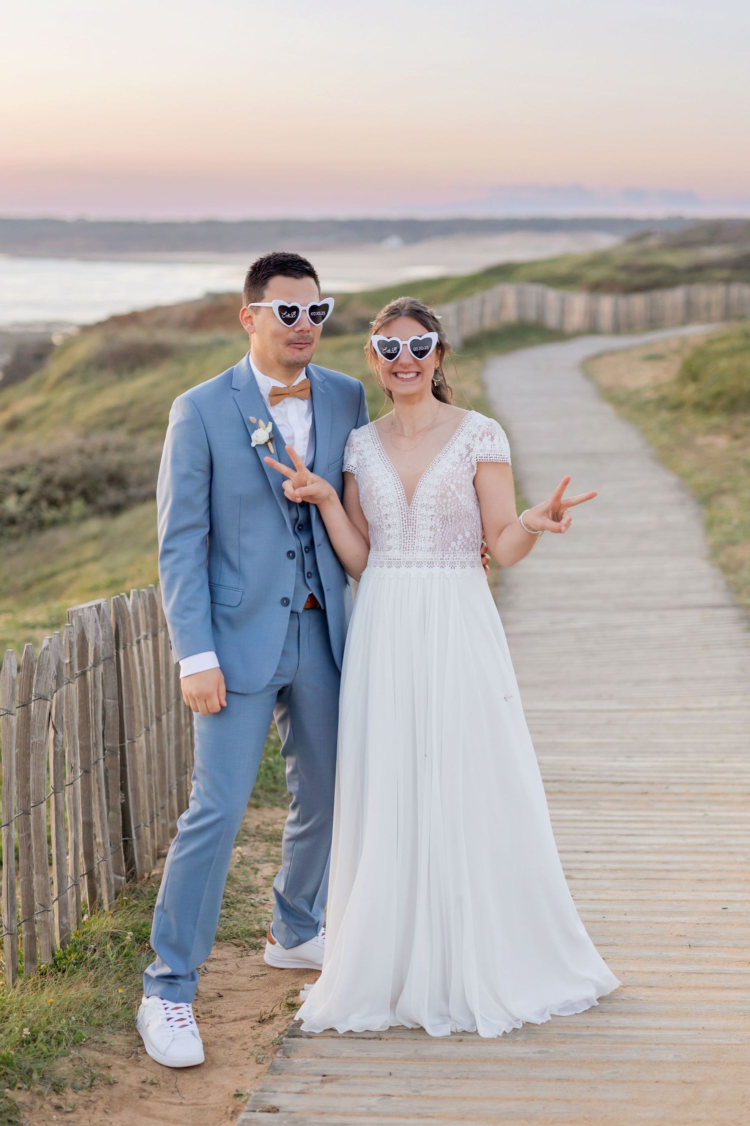 Couple in wedding attire posing on a coastal boardwalk with blue suit and white dress during sunset.
